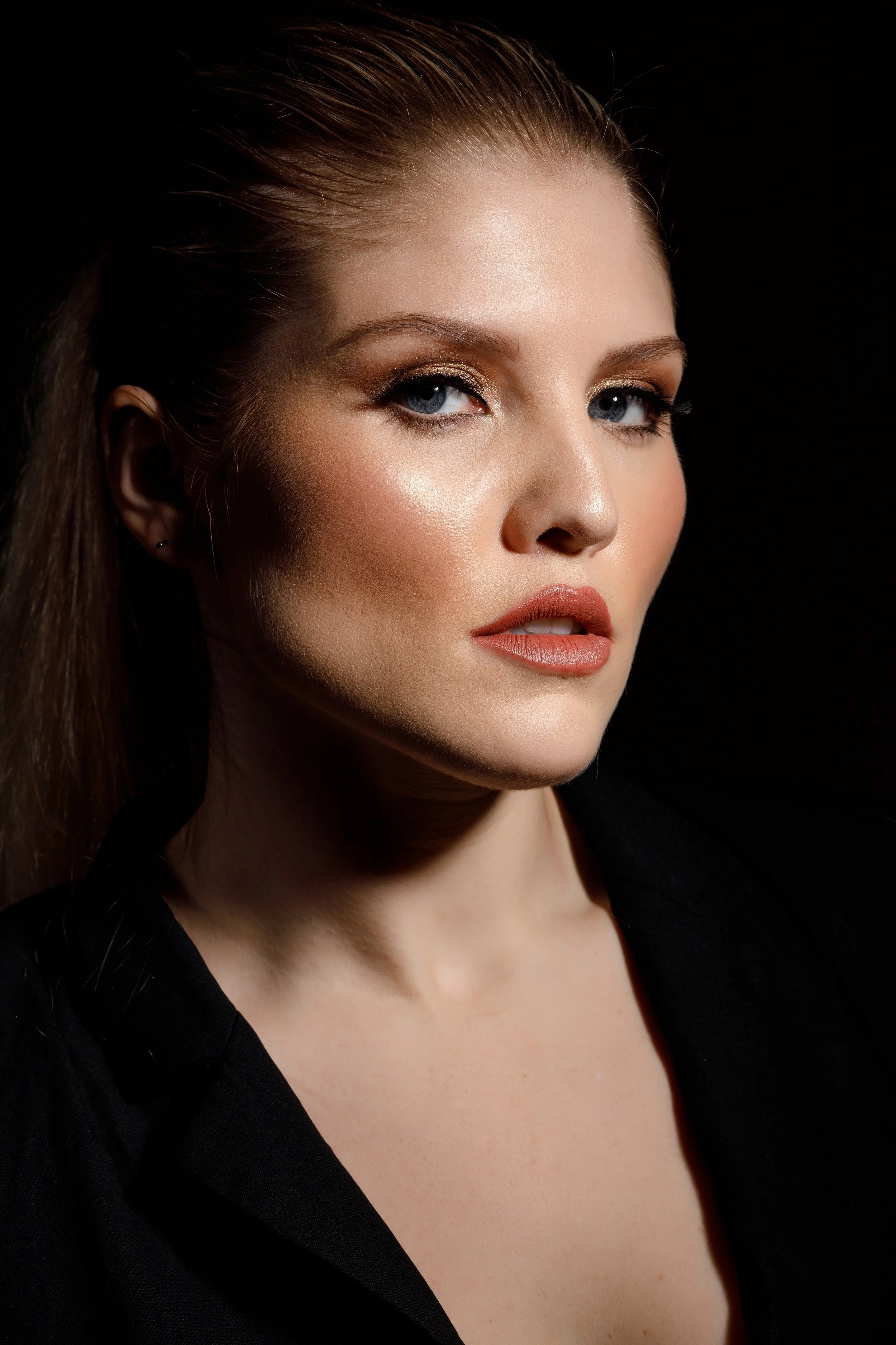 Close-up portrait of a woman with blue eyes, fair skin, and brown hair slicked back, wearing makeup with gold eyeshadow and peach lipstick, dressed in a black top against a black background.