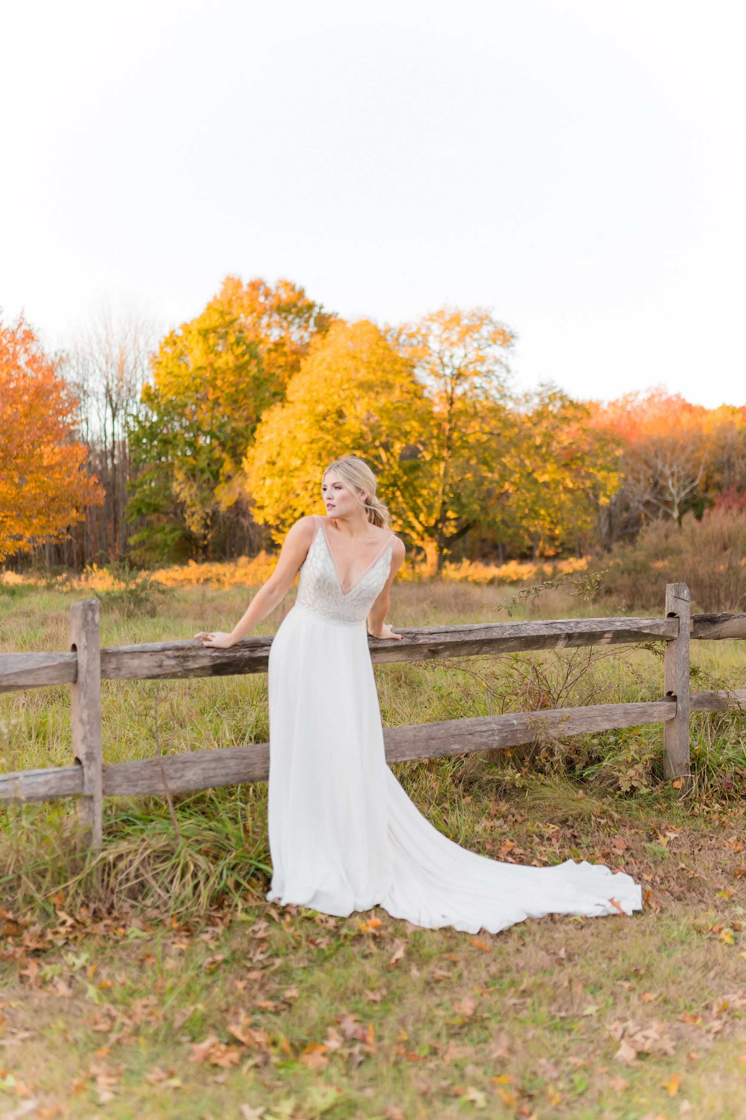 A woman in a white dress leaning on a wooden fence in a field with autumn trees with orange, yellow, and red leaves in the background.