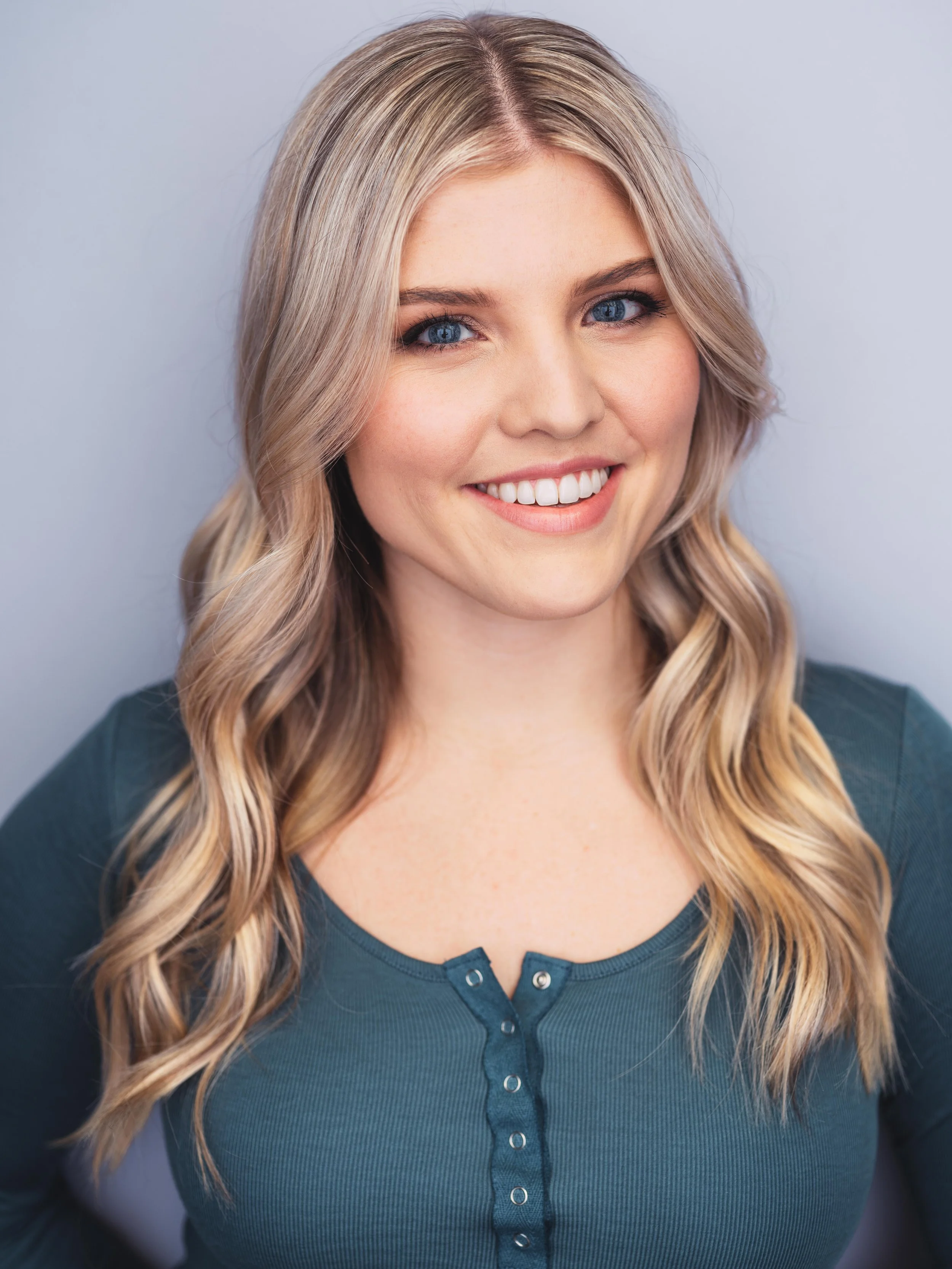 A woman with blonde wavy hair and blue eyes smiling, wearing a dark teal top with buttons, against a plain light gray background.