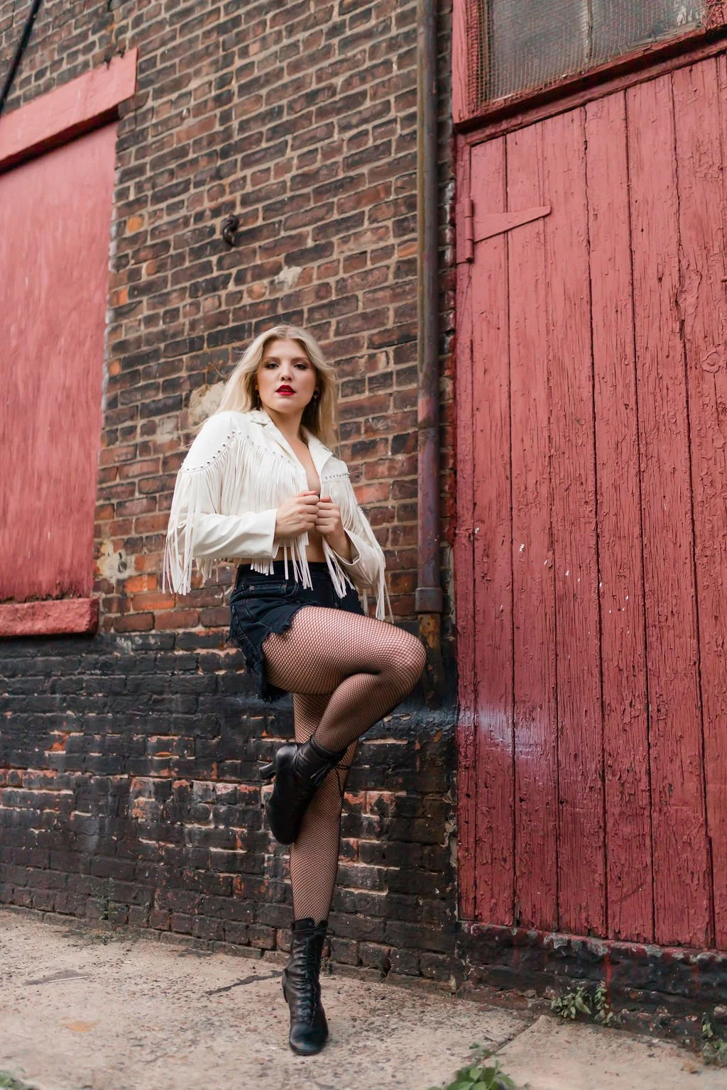 A woman with blonde hair wearing a cream-colored fringed jacket, black shorts, fishnet tights, and black combat boots, poses against a brick alley wall adorned with red wooden doors.