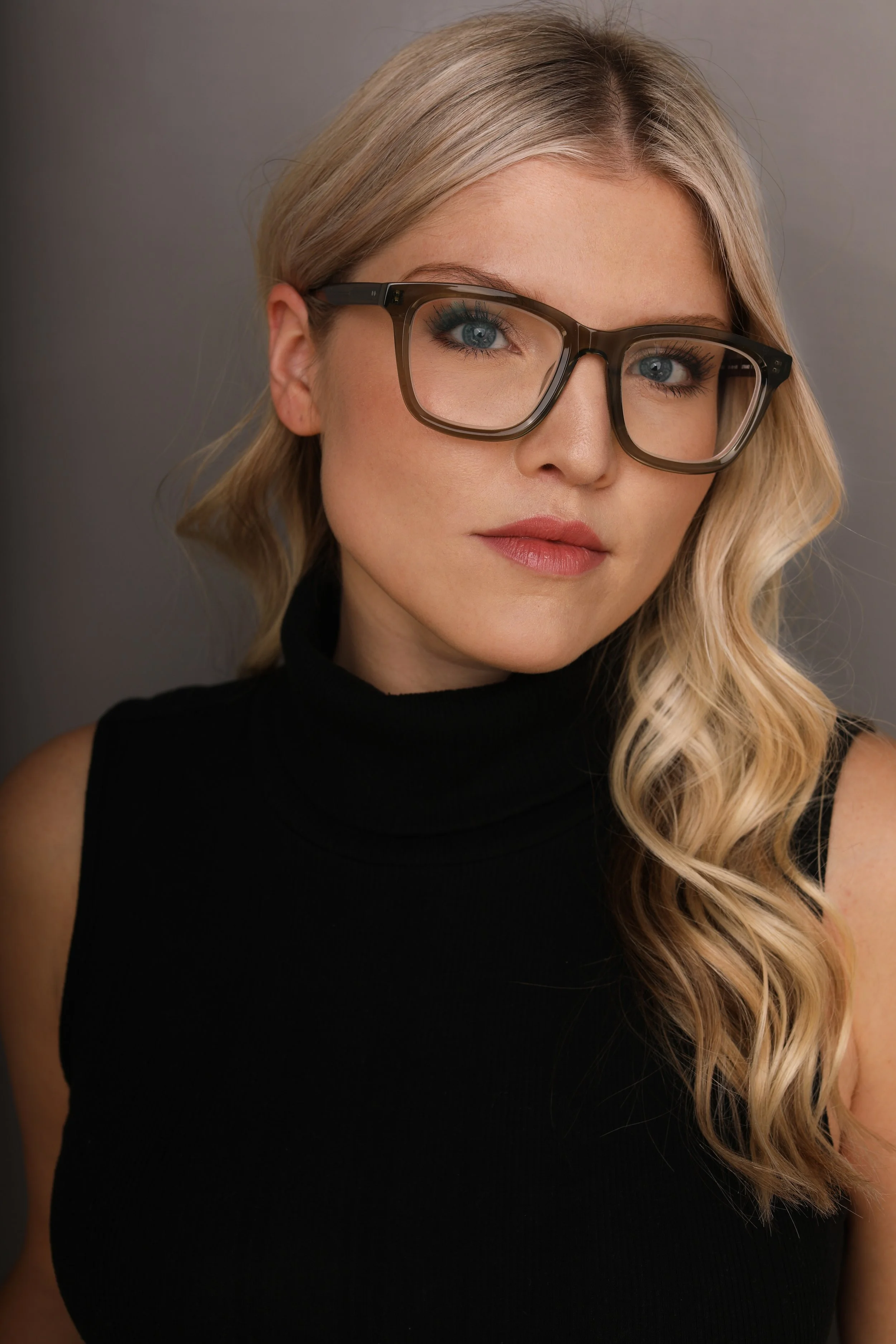 Close-up portrait of a woman with long wavy blonde hair wearing large black glasses and a black sleeveless turtleneck top, looking directly at the camera with a slight smile.