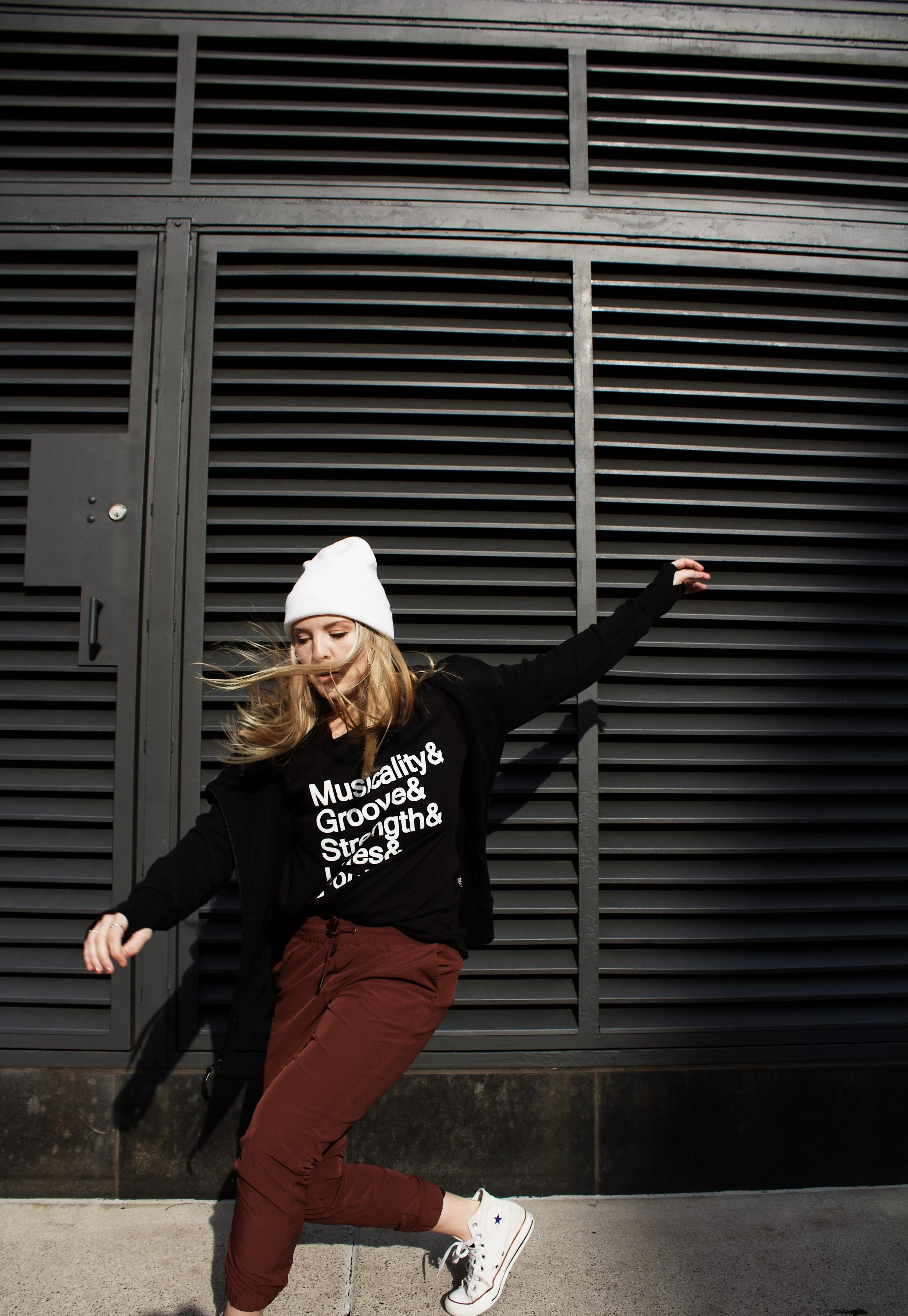 A young woman wearing a white beanie, black sweatshirt with white text, maroon pants, and white sneakers, dancing or jumping in front of a modern black metal wall with horizontal slats.