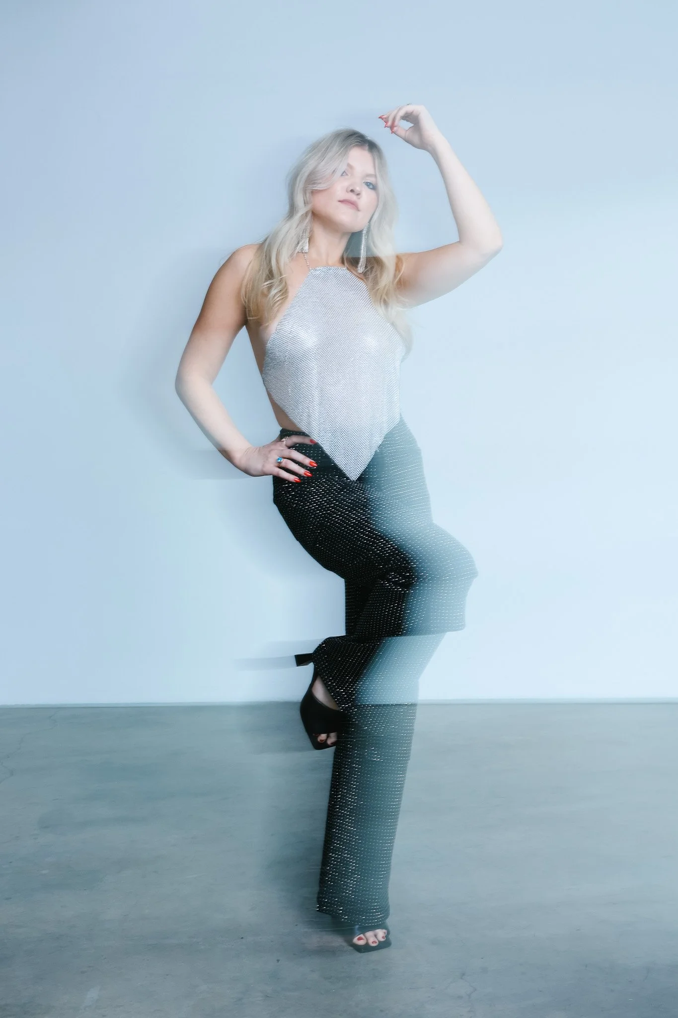 A woman with blonde hair wearing a sparkly sleeveless top and black polka dot pants, standing against a plain light-colored wall, with one hand on her hip and the other raised above her head.
