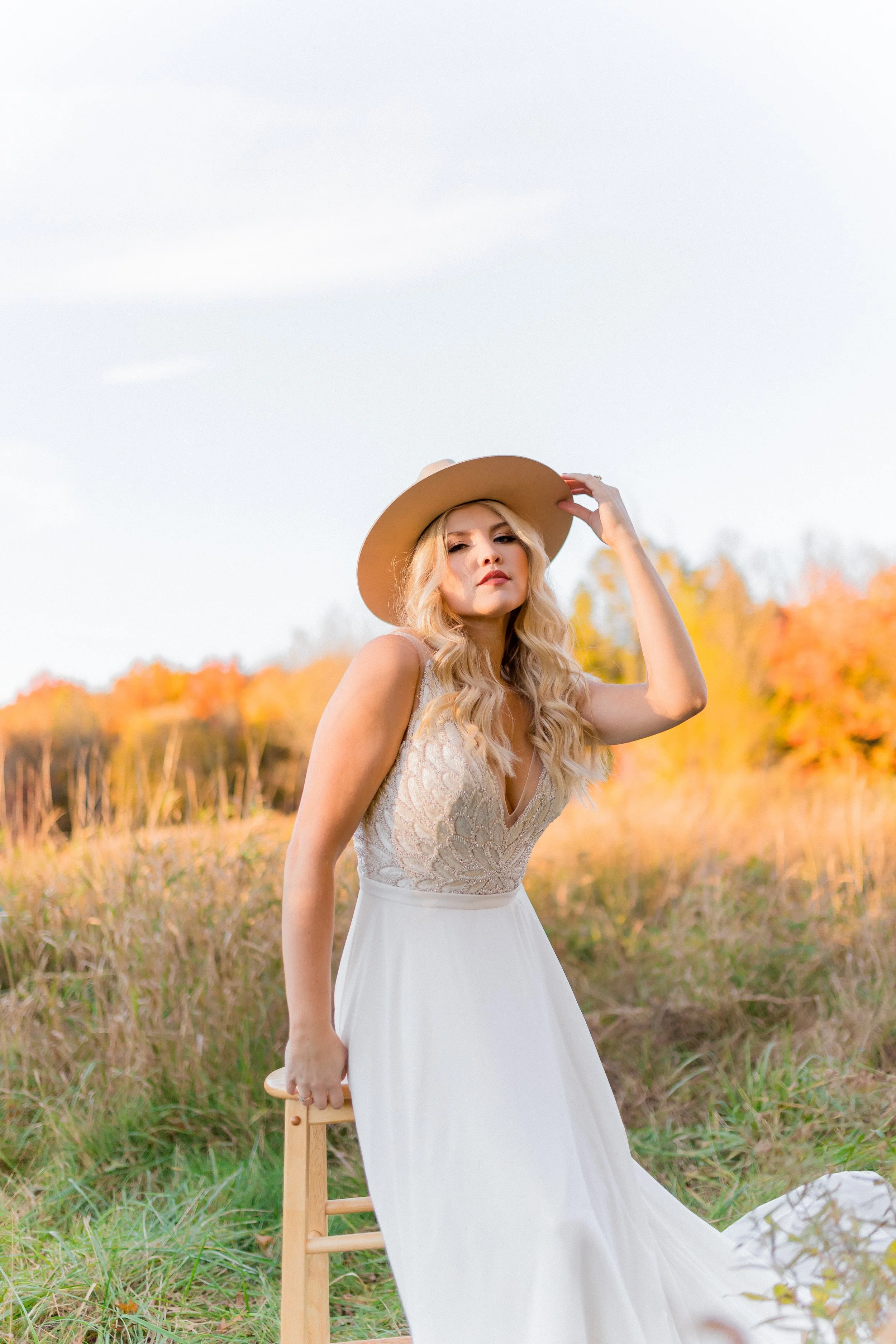 A woman with long blonde hair wearing a white lace dress and wide-brimmed hat standing outdoors in a field with autumn-colored trees in the background.