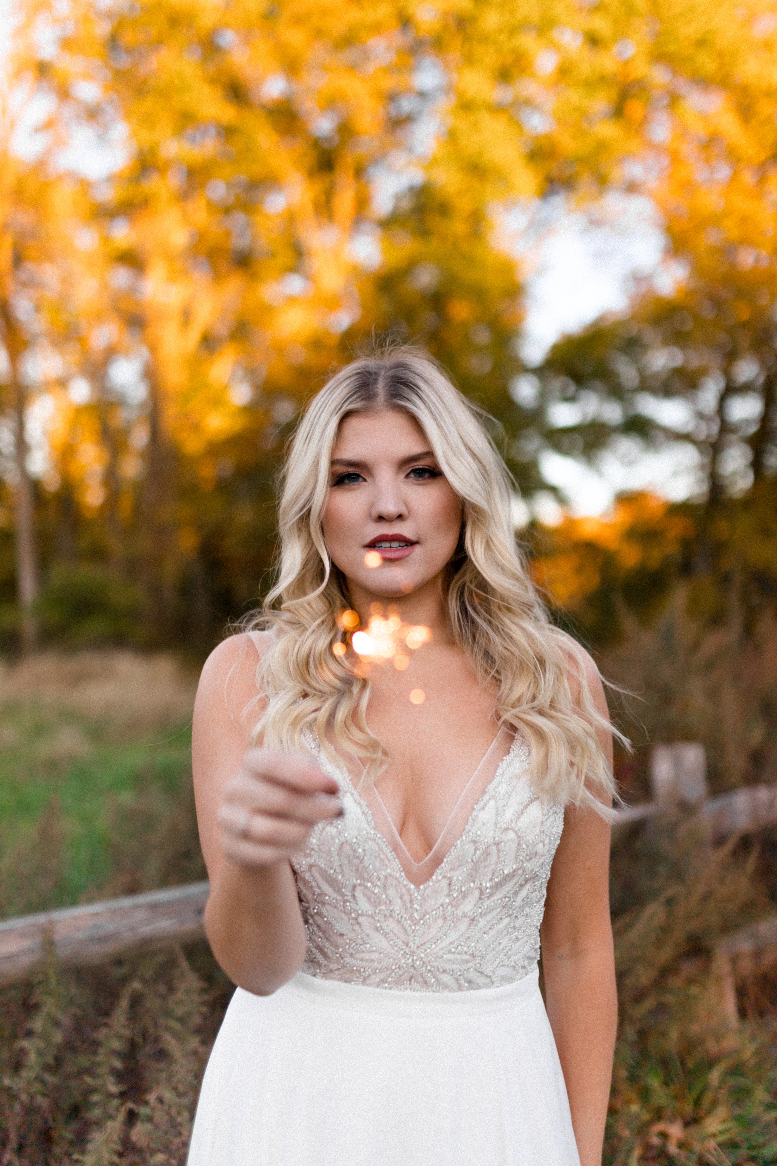 A woman in a white lace dress standing outdoors during fall, with yellow-orange trees in the background, holding a sparkler.