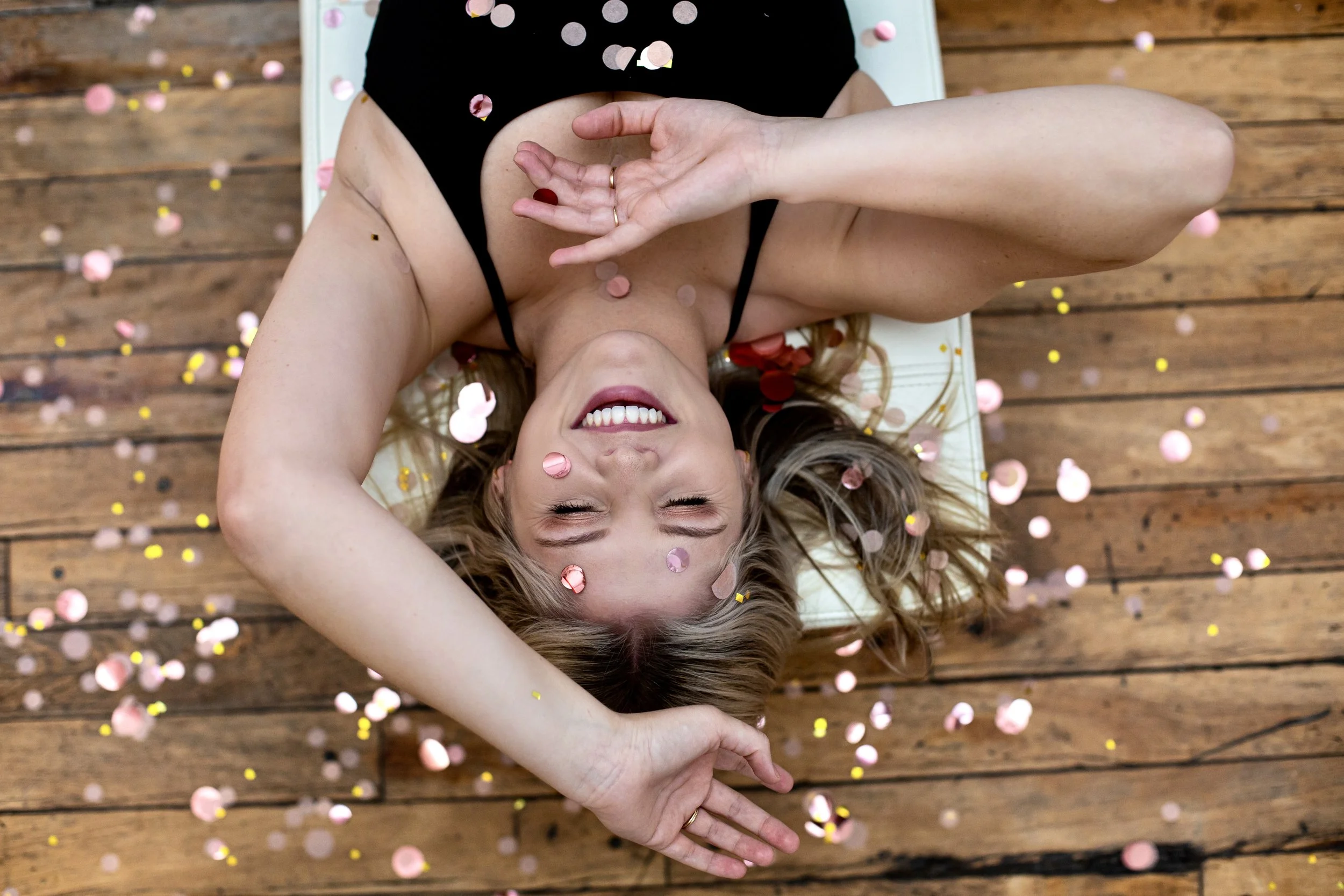 A woman lying on a wooden floor covered with pink and yellow confetti, smiling, with her arms behind her head, wearing a black dress.