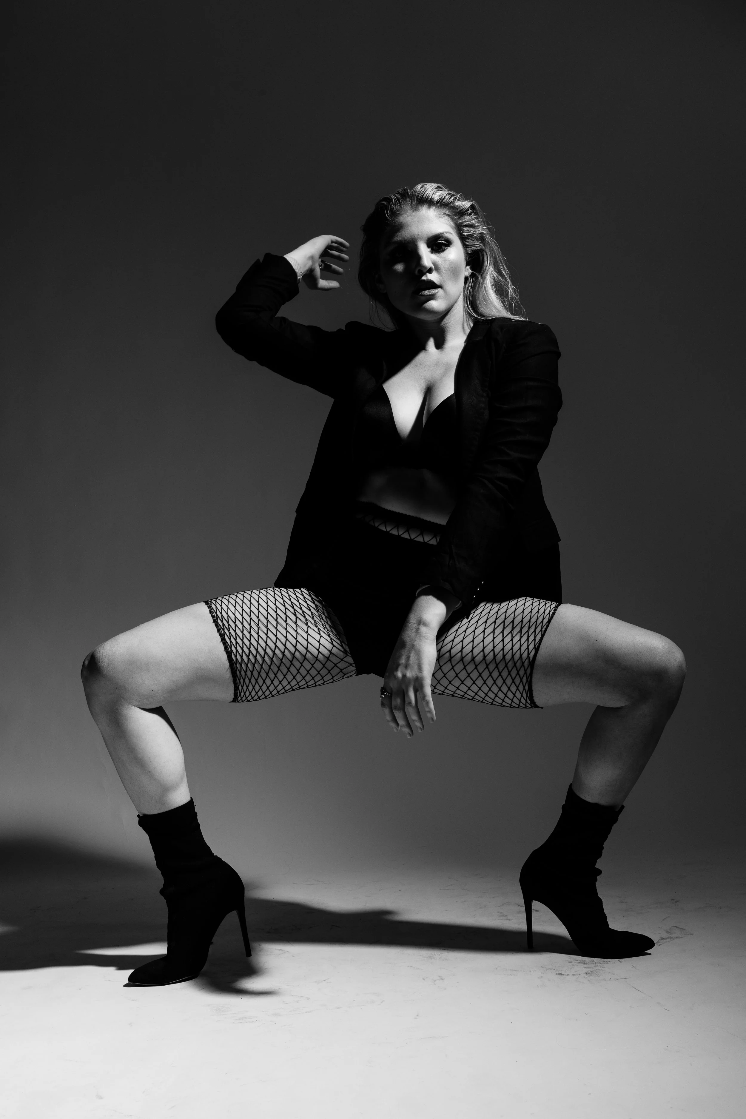 Black and white photo of a woman in a provocative pose, wearing fishnet stockings, high heels, and a revealing outfit, with her legs spread wide, one arm behind her head, and the other hand resting on her ankle.