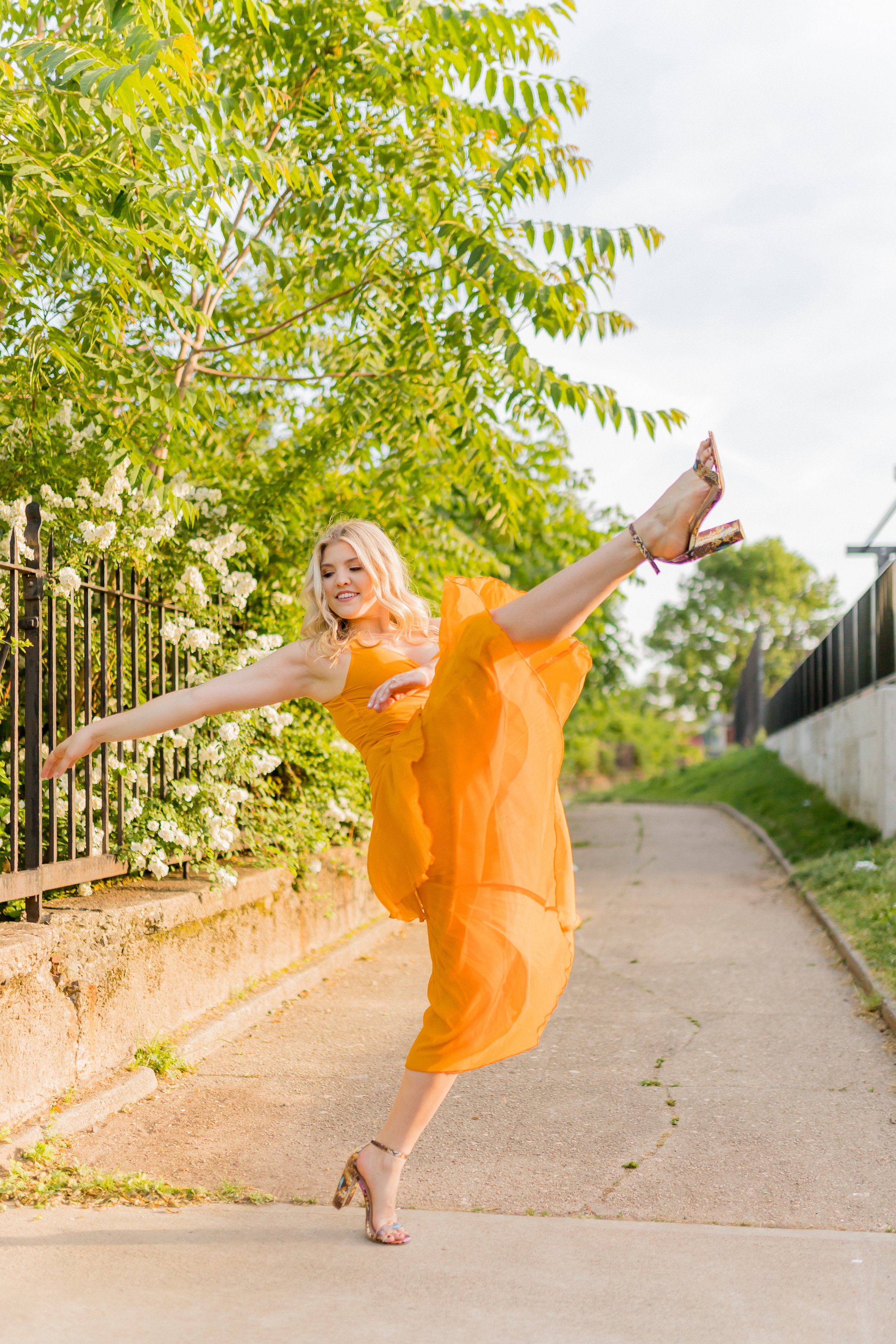 A woman with blonde hair wearing an orange dress and high heels is performing a high kick outdoors on a sidewalk.
