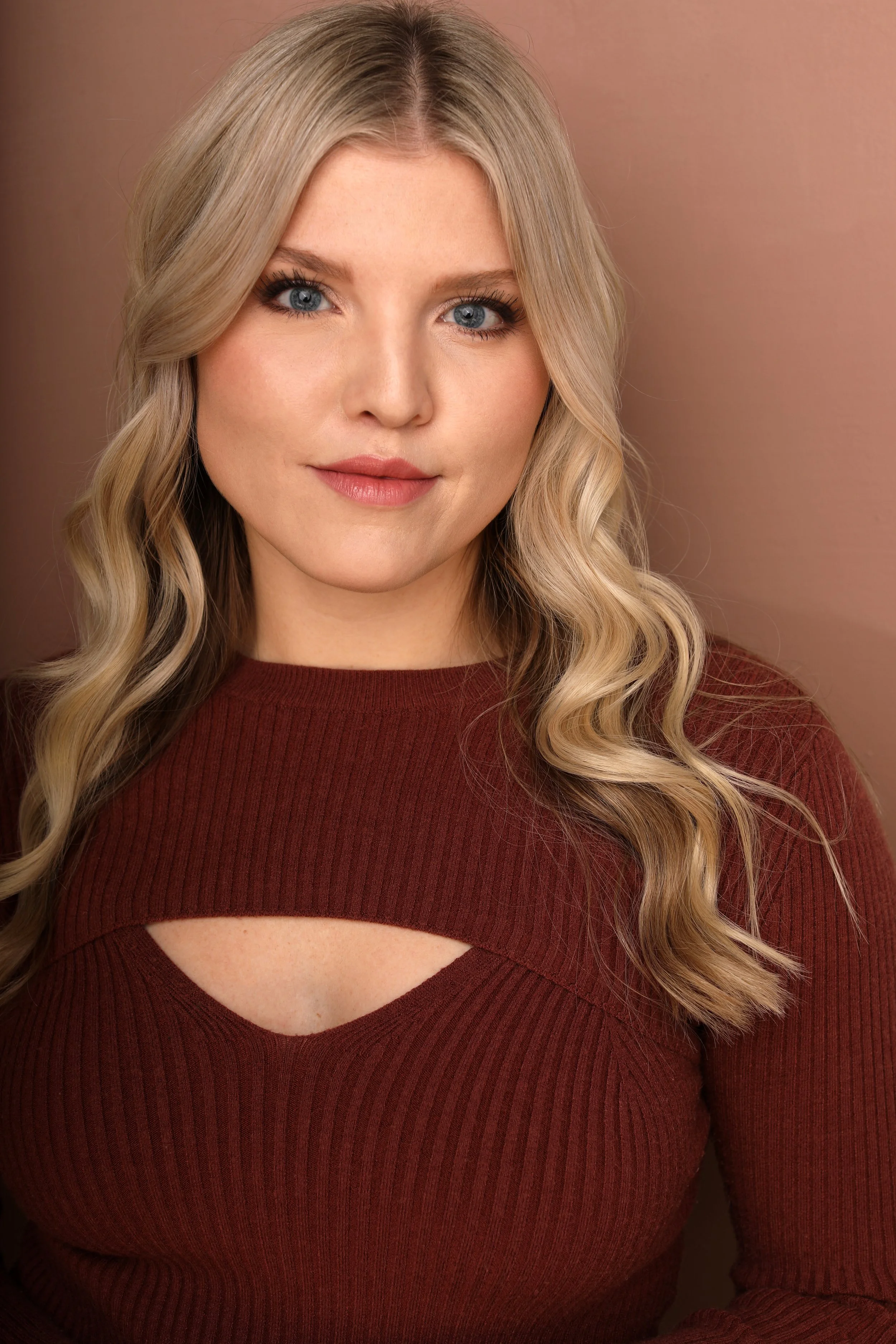 A woman with long blonde hair, blue eyes, wearing a maroon top with a cut-out design, standing in front of a plain beige background.