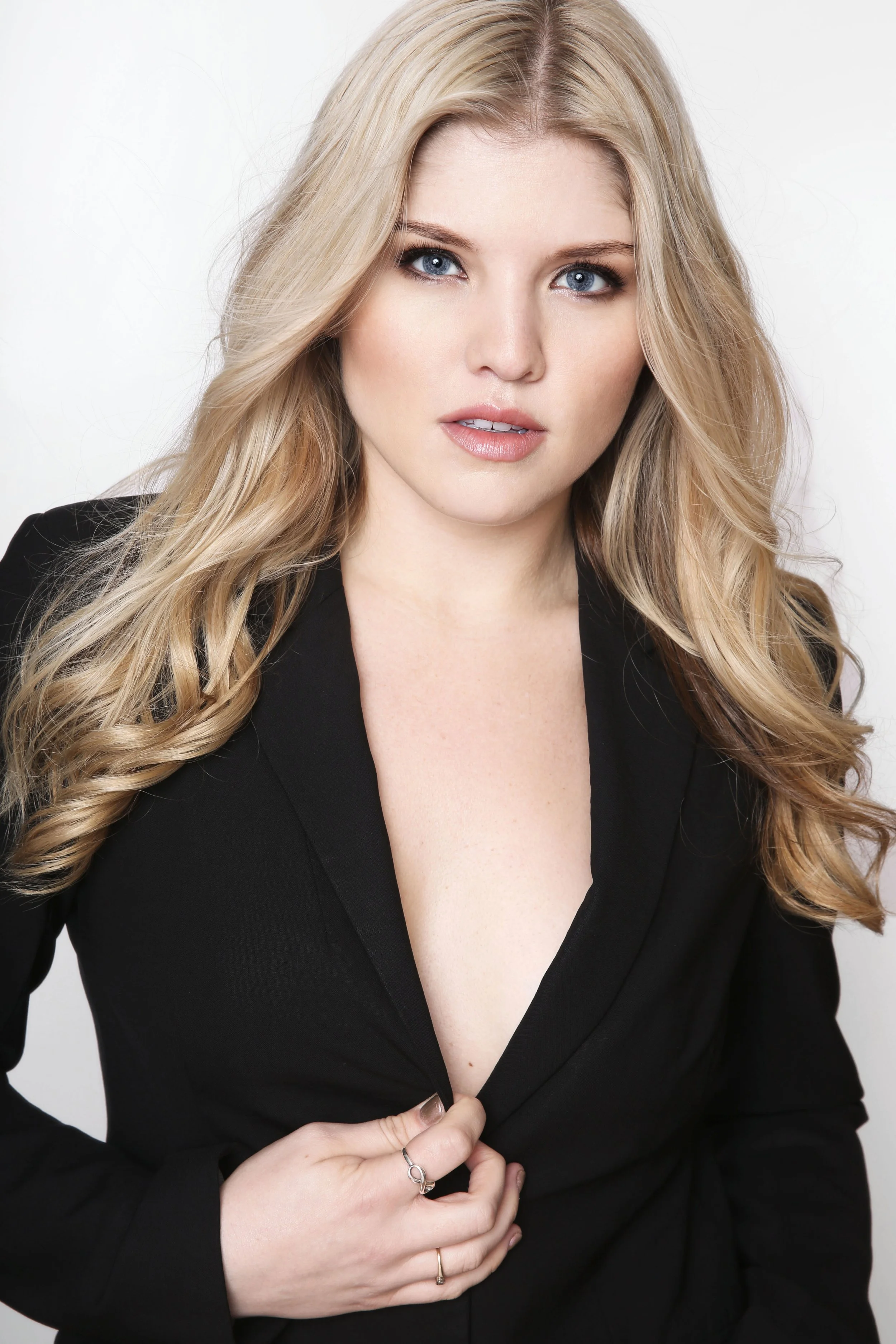 A woman with long, wavy blonde hair and blue eyes wearing a black blazer and jewelry, touching the button of her blazer with her left hand, against a plain white background.