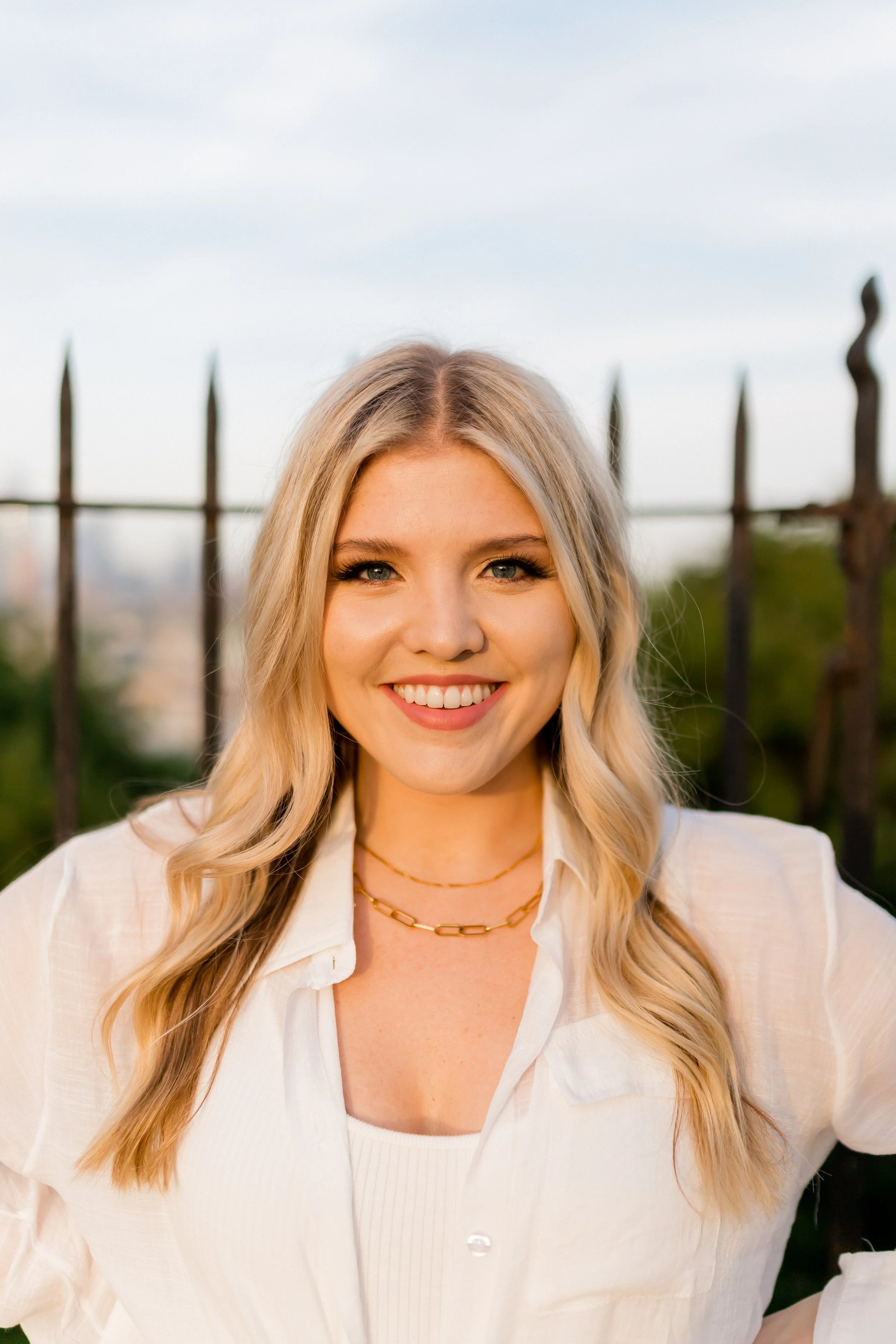 Close-up of a young woman with blonde wavy hair, smiling, wearing a white blouse and gold jewelry, standing outdoors near a black metal fence with a blue sky in the background.