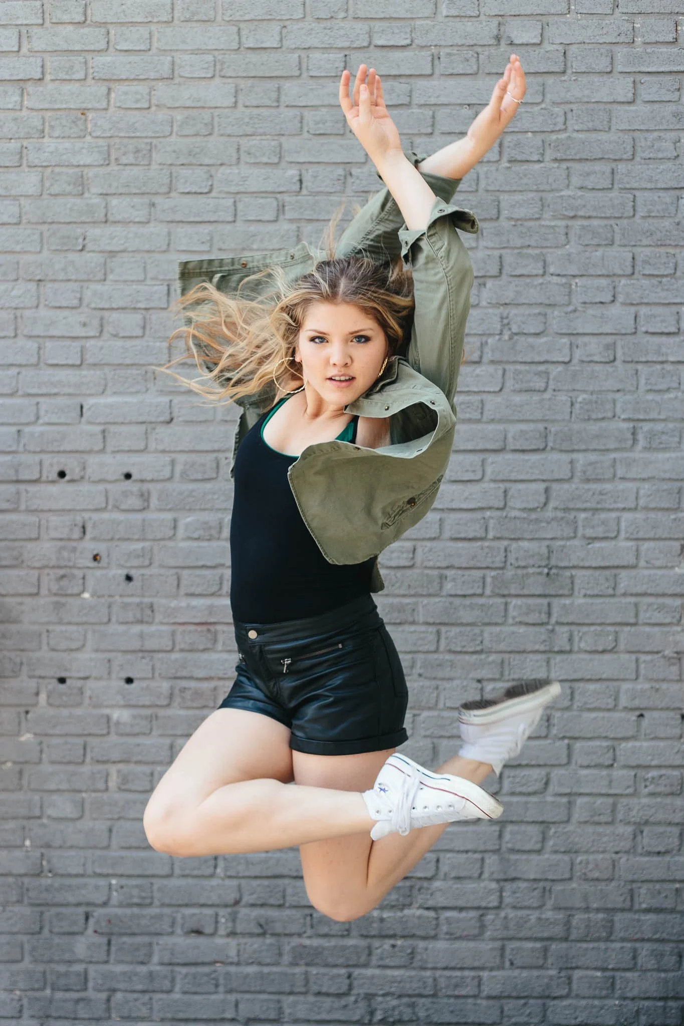 A young woman with wavy blonde hair and blue eyes poses in front of a gray brick wall. She wears a black tank top, black shorts, a green jacket, and white sneakers. She appears to be jumping or dancing with a confident expression.