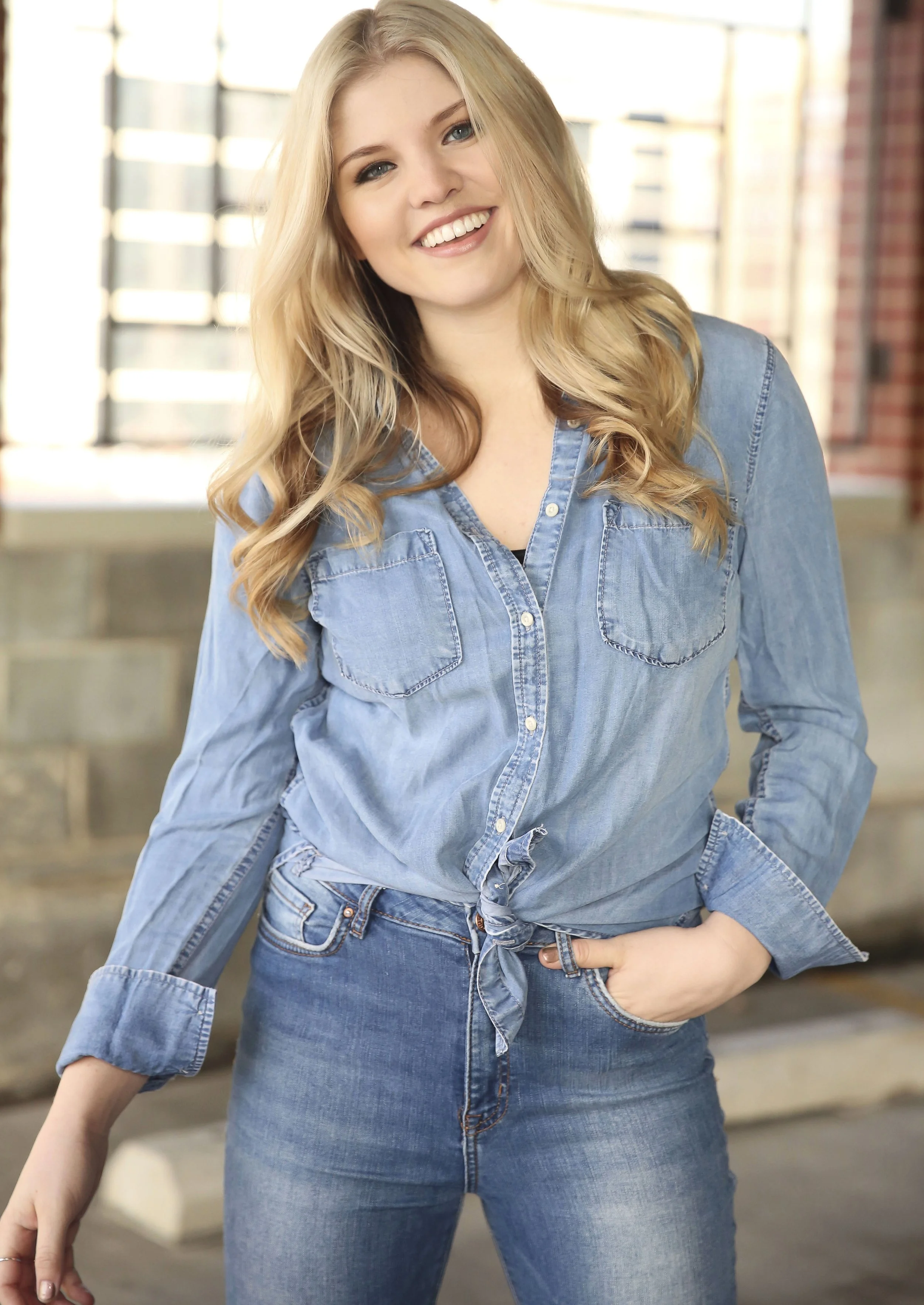A smiling young woman with long blonde hair wearing a denim shirt and jeans, standing indoors near large windows with blurred brick walls in the background.