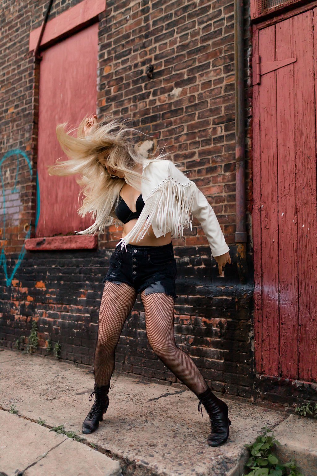 A woman dancing with her hair in motion, wearing a black top, distressed black shorts, fishnet stockings, a white fringed jacket, and black boots, standing on a sidewalk against a brick wall with red and purple wooden panels.