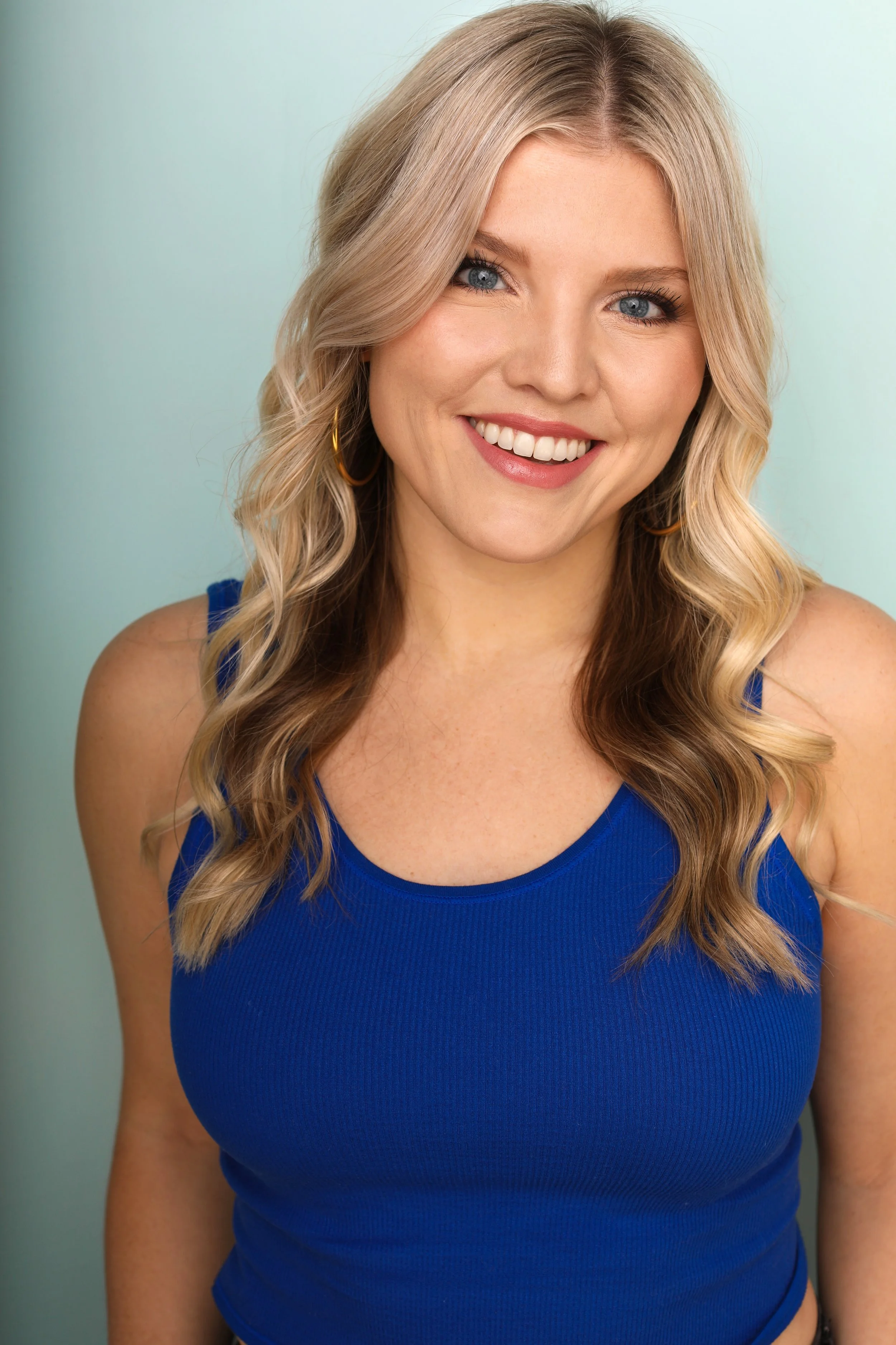 Close-up portrait of a smiling young woman with long wavy blonde hair wearing a royal blue sleeveless top and gold hoop earrings against a light green background.