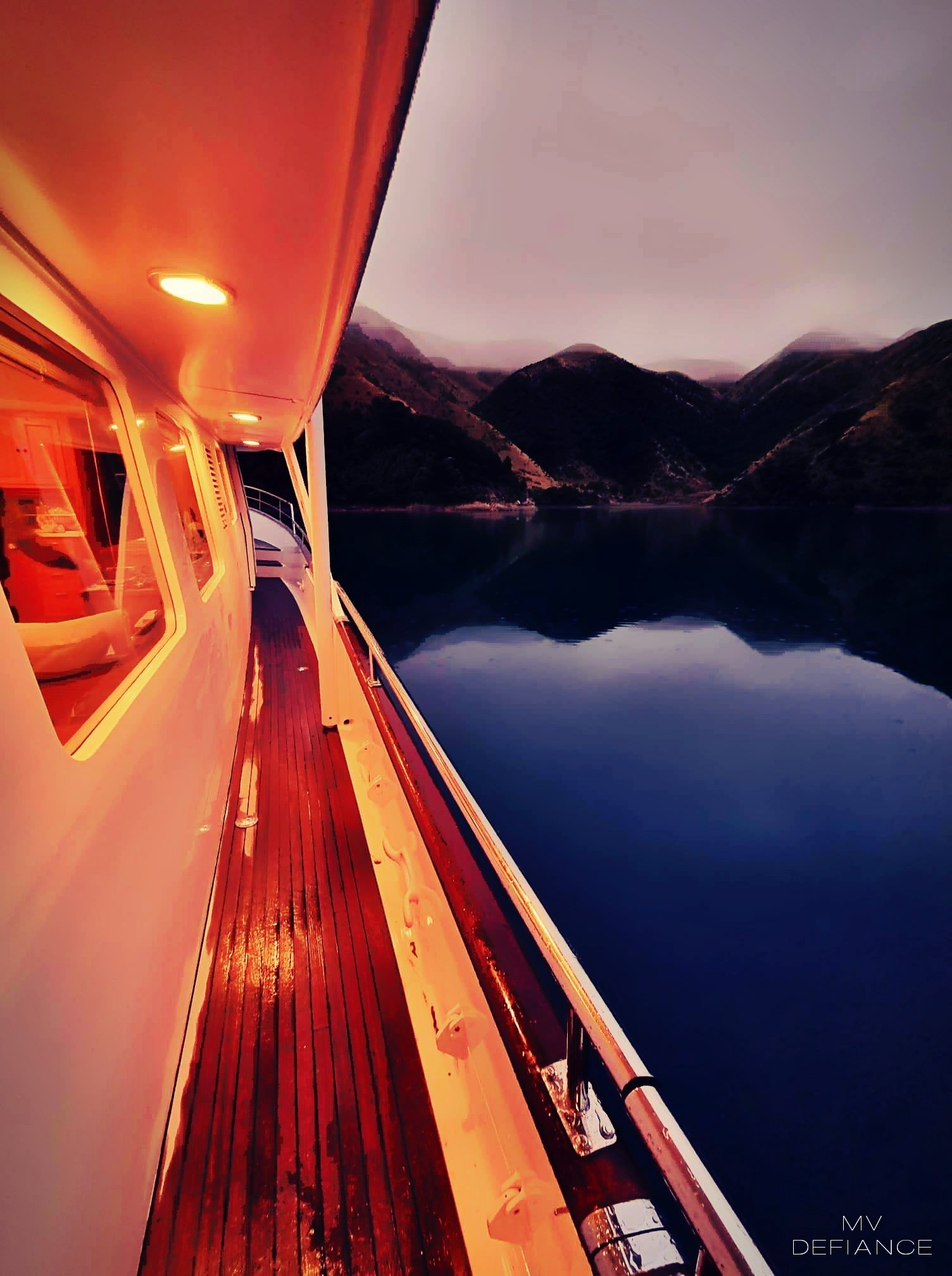 The side view of a charter boat in the Marlborough Sounds deck with a warm orange glow from the ceiling lights, sailing on calm water with mountain hills in the background during sunset or sunrise.