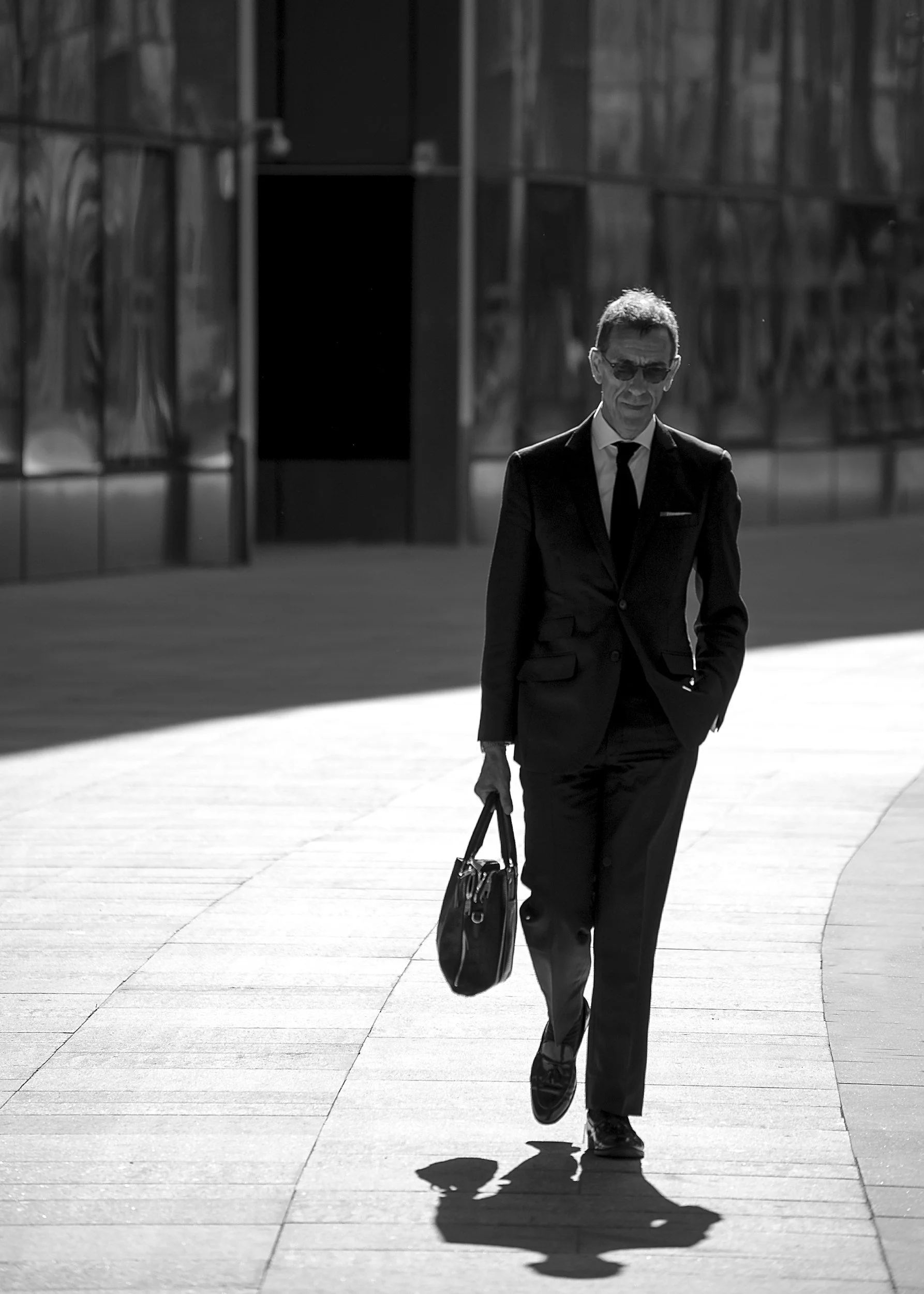 Black and white photo of a man in a suit walking outside on a sunny day, carrying a bag.