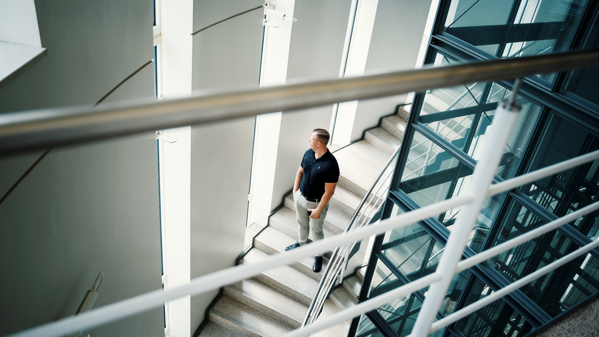 A man standing on a staircase near large windows in a modern building, looking out.