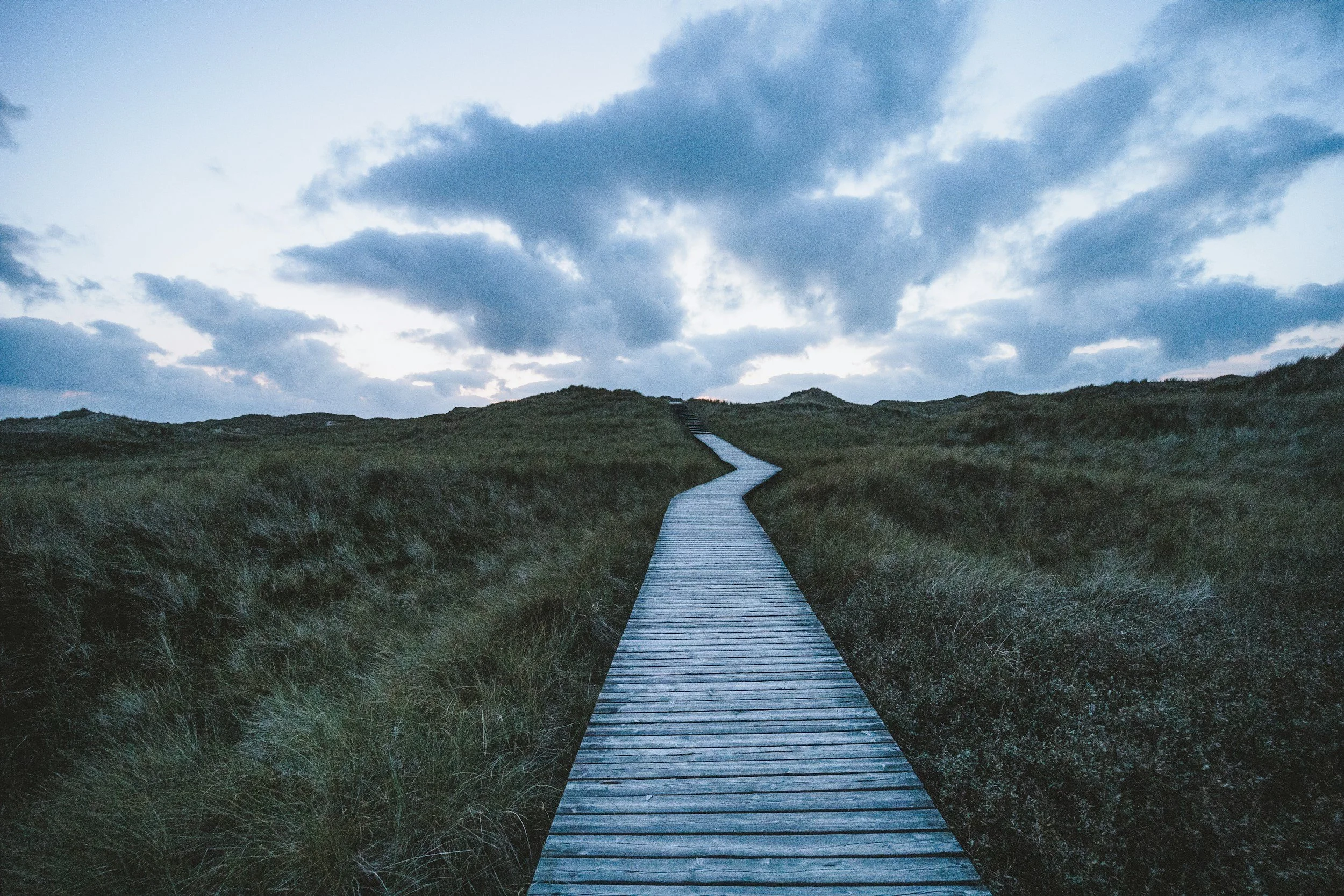 A wooden boardwalk winding through grassy dunes under a cloudy sky.