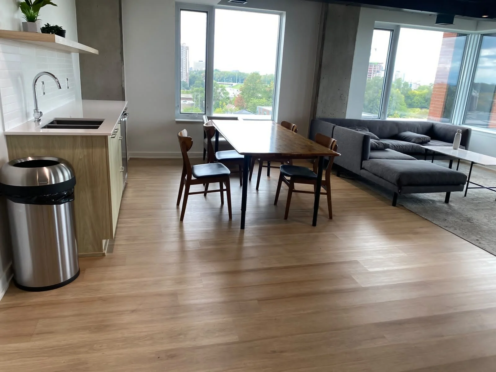 Modern apartment interior with a kitchenette, wooden dining table with four chairs, gray sectional sofa, large windows showing cityscape, and hardwood floors.