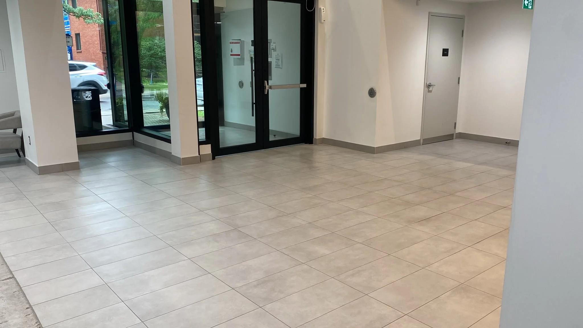 Interior of a building lobby with beige tiled floor, glass doors leading outside, and white walls. A green exit sign is visible at the top right corner. Outside the glass doors, a white car and some greenery are visible.