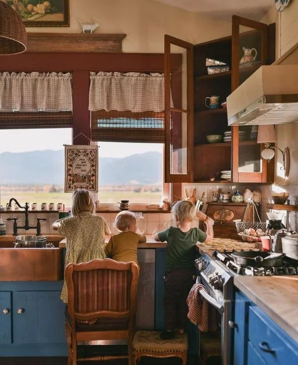 Three children at a rustic kitchen counter looking out the window at a rural landscape.