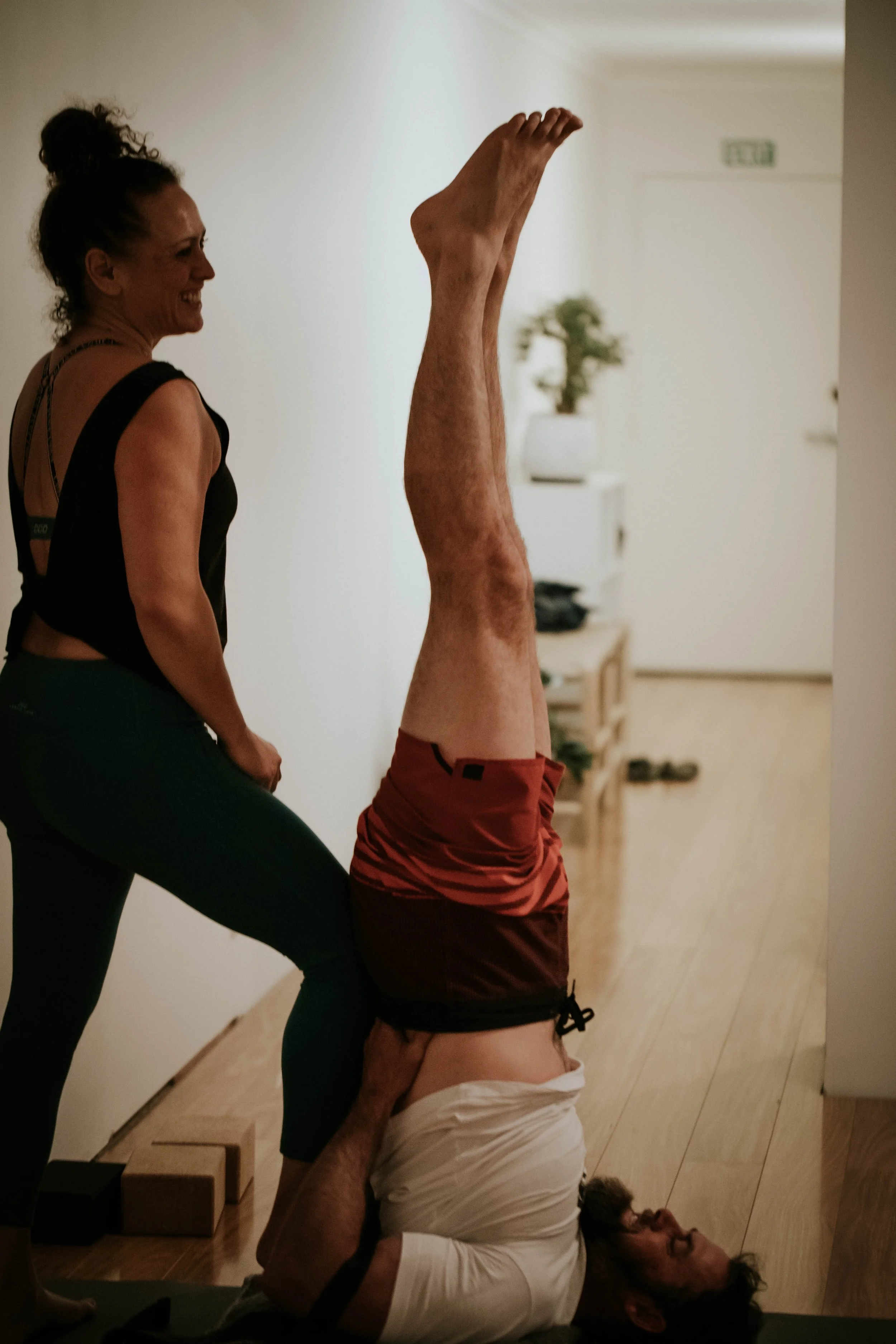 Group of people practicing yoga indoors, performing various poses including headstands and arm balances, in a well-lit room with wooden flooring and a potted plant.