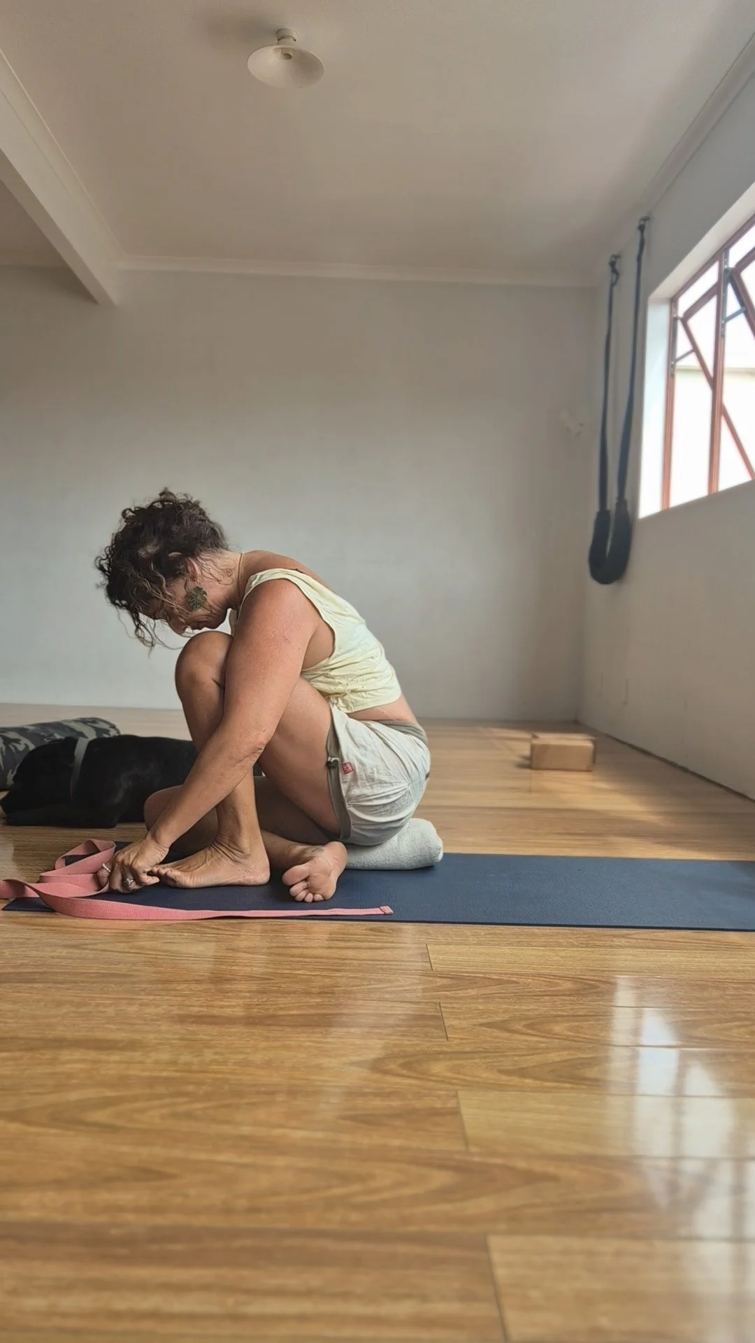 A person with curly hair and tattoos is practicing yoga on a mat, sitting in a curled position and tying their pink yoga strap, with a black dog resting nearby in a minimalist room with a wooden floor and a window.