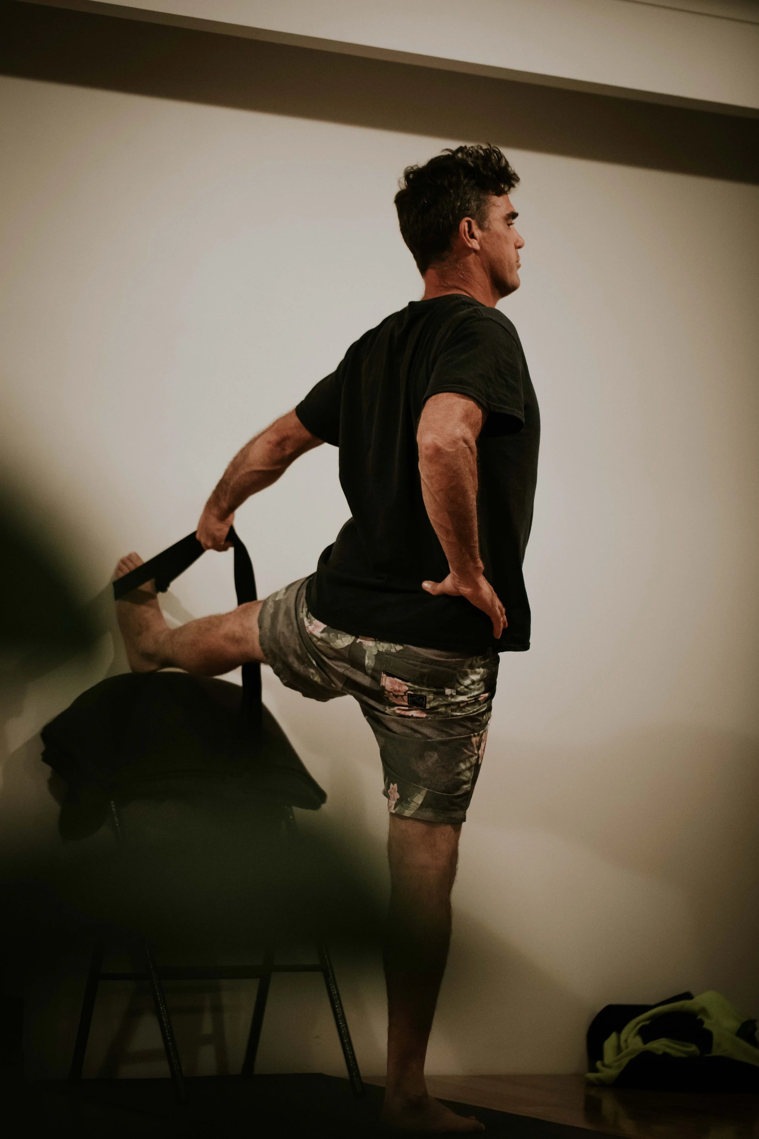 A man practicing yoga indoors with one leg bent on a chair, supporting himself with his hand on his waist, wearing a black t-shirt and camouflage shorts.