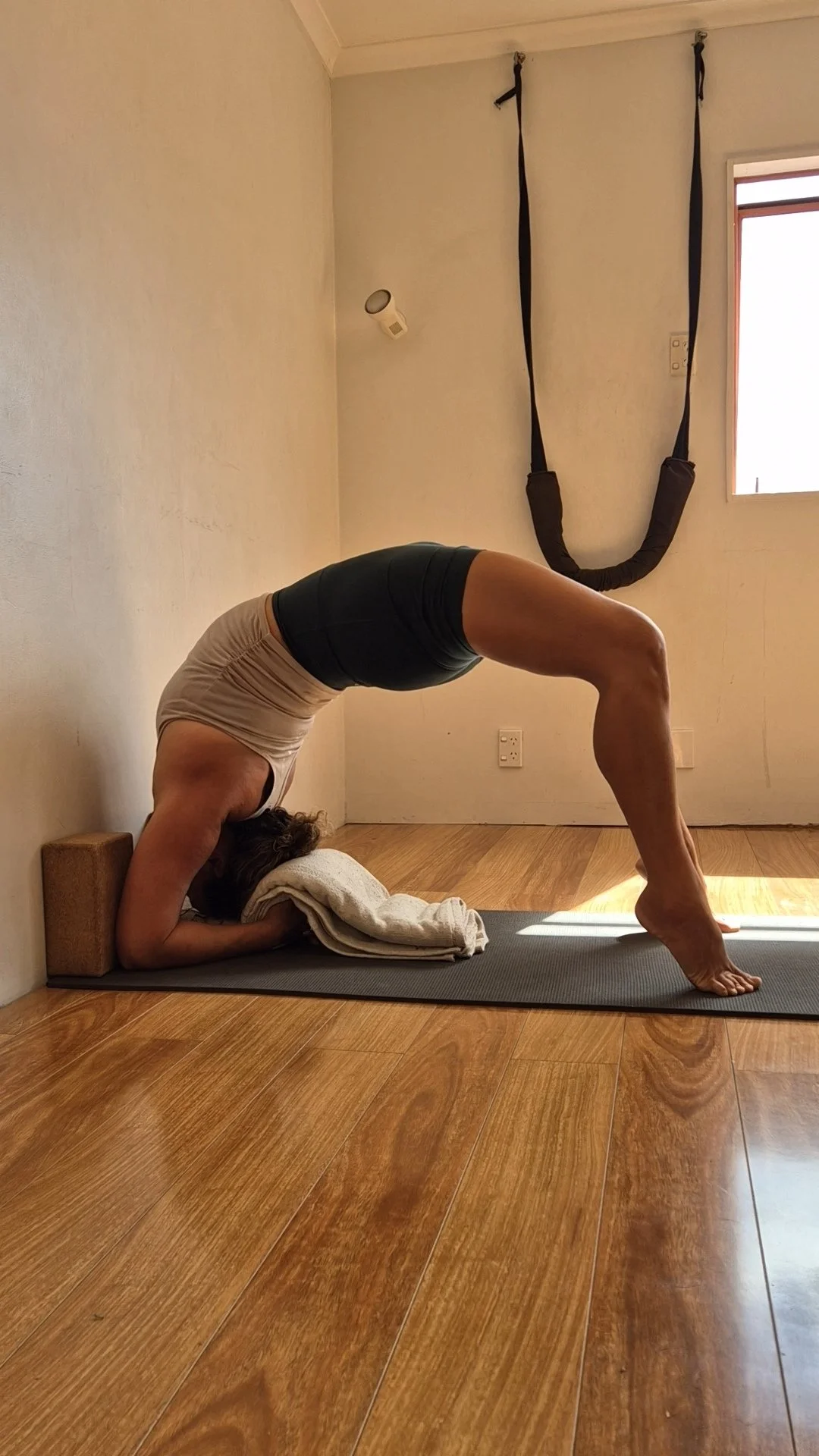 Woman practicing yoga in a downward-facing dog pose with her hands on a folded towel, head on a cushion, in a well-lit room with wooden flooring, an exercise strap hanging from the ceiling, and a window.