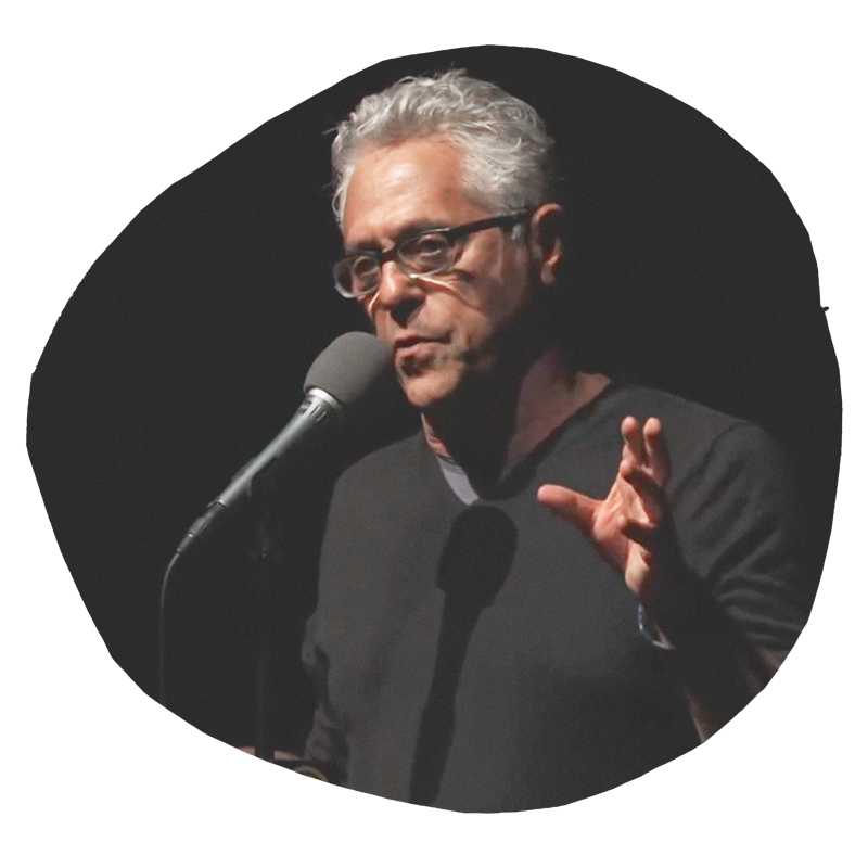 A man with gray hair and glasses speaking into a microphone on a dark stage.
