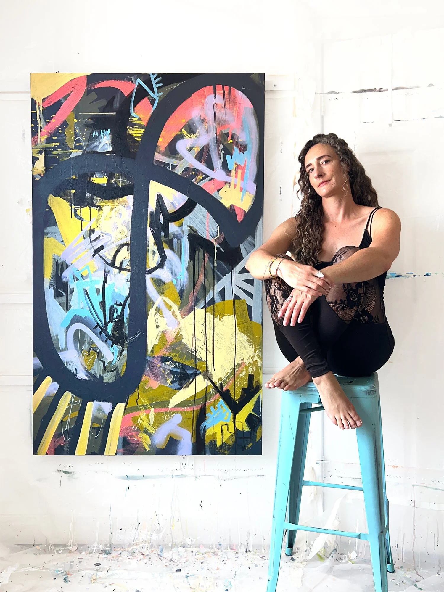 The artist, Marissa Caputo's studio, with an abstract painting on the wall, she's sitting on a blue stool next to it. She is dressed in black lace clothing, with one knee pulled up and her arms crossed over it, smiling at the camera.