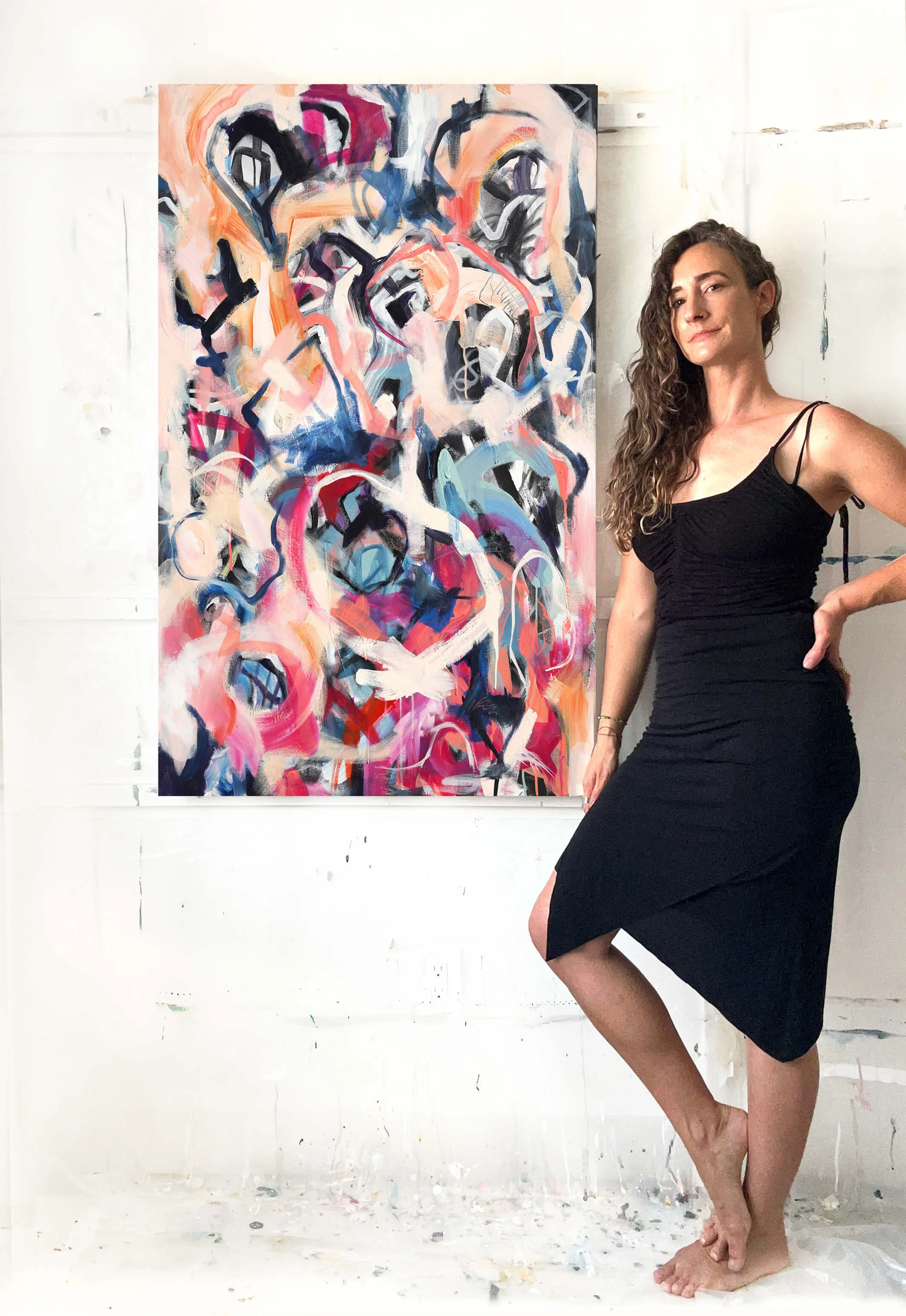 Marissa Caputo, a woman, wearing a black dress poses next to her abstract colorful painting on a white wall. The floor and wall are splattered with paint.