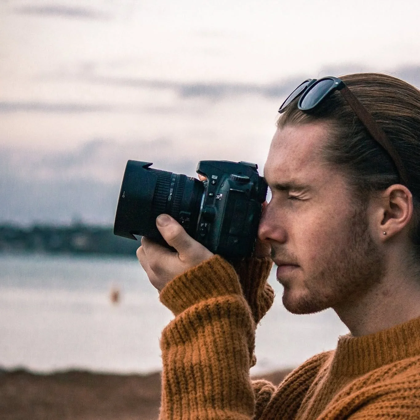A young man with styled hair, wearing sunglasses on his head, a mustard-brown sweater, and earrings, is taking a photograph with a DSLR camera near the seaside at sunset or early evening.