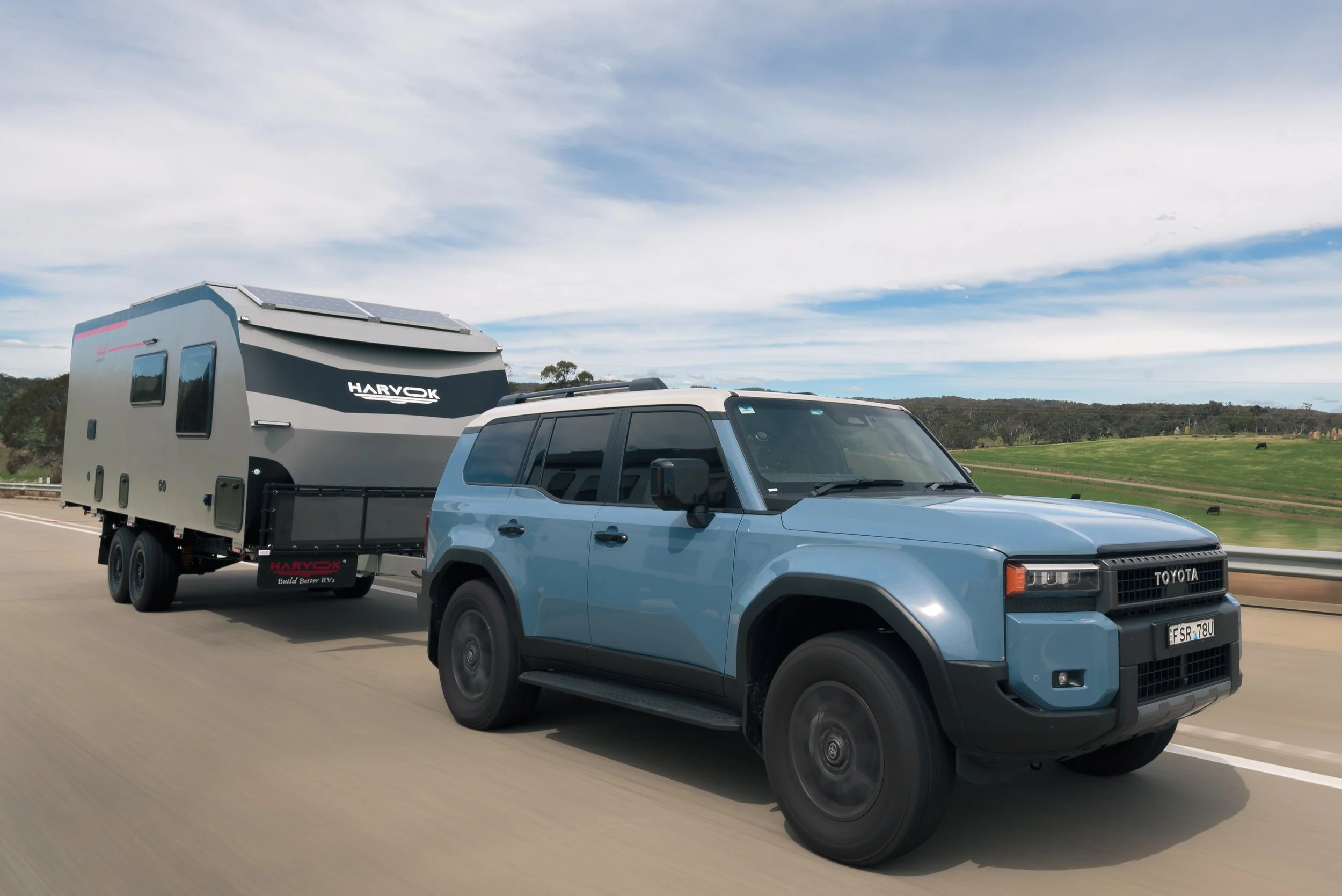 Blue Toyota truck towing a travel trailer on a highway with grassy fields and trees in the background under a partly cloudy sky.