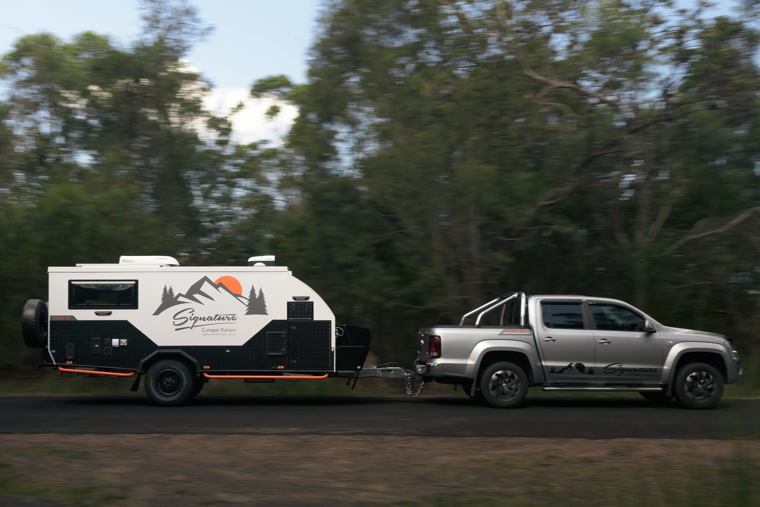 A pickup truck towing a travel trailer with a forested background and movement blur indicating motion.