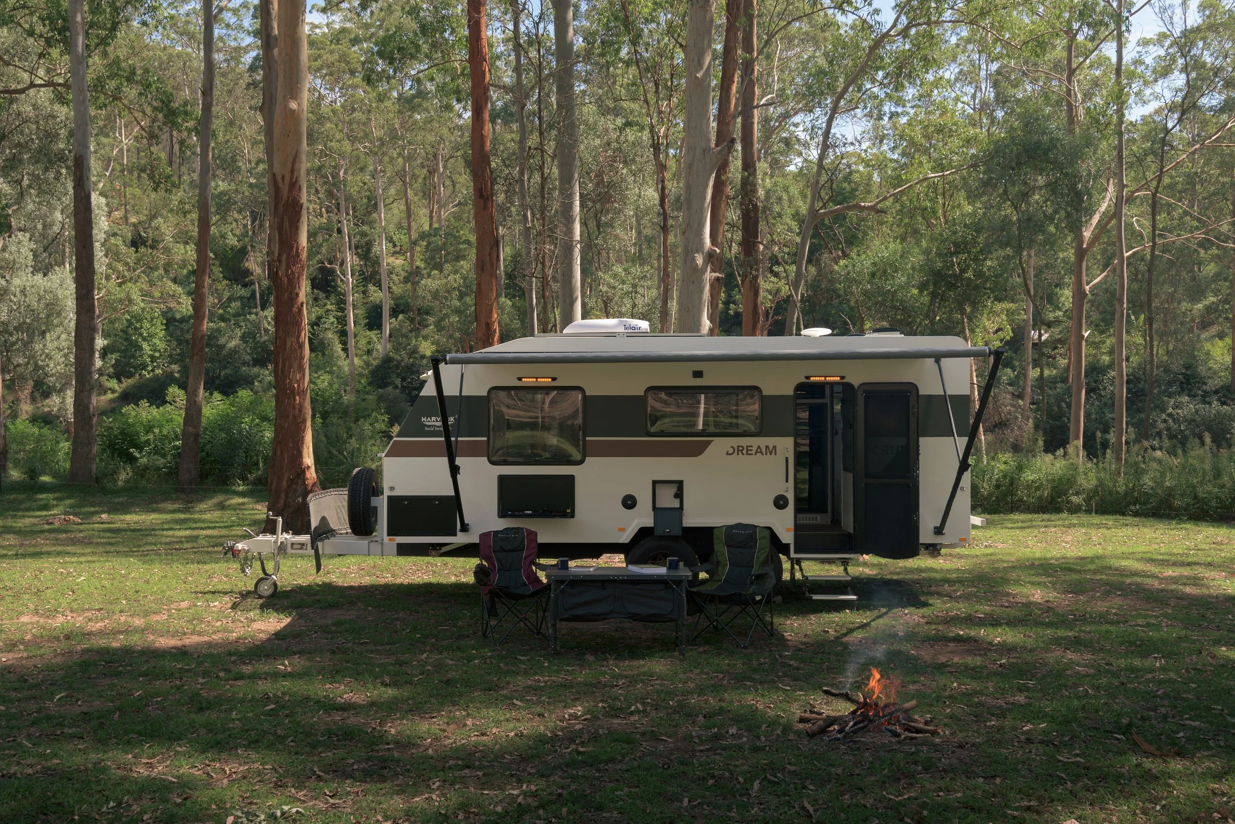A camper trailer parked on a grassy area surrounded by tall trees in a forest, with a small campfire burning in the foreground.