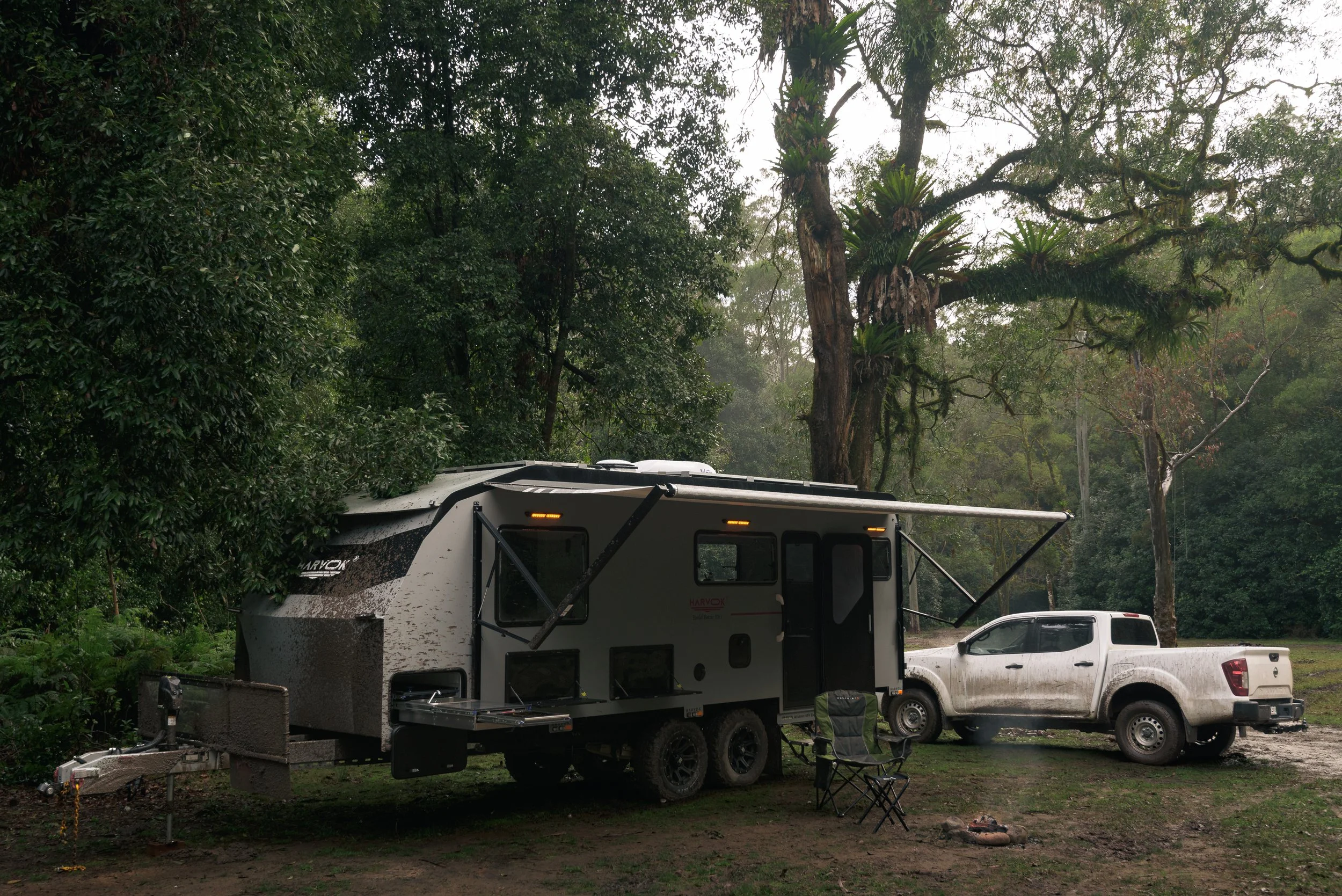 A rv trailer with an open hatch in a wooded campground, with a metal chair and a white pickup truck nearby.