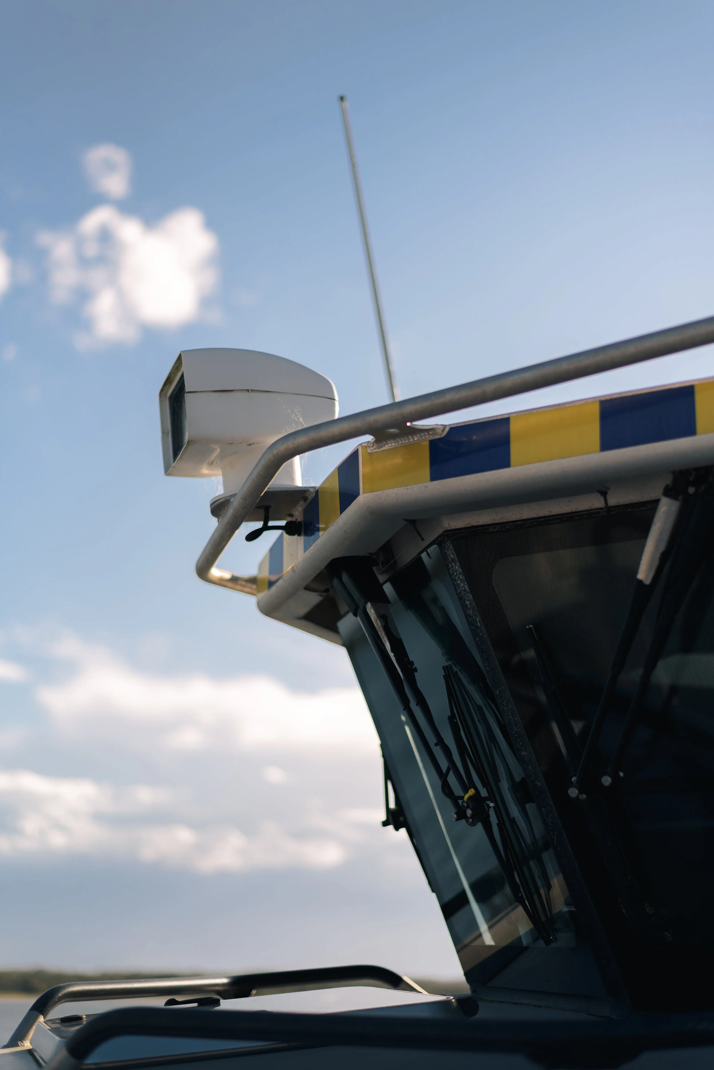 A close-up view of a road vehicle's roof with various mounted equipment, including a white weather sensor and a tall antenna, against a blue sky with scattered clouds.