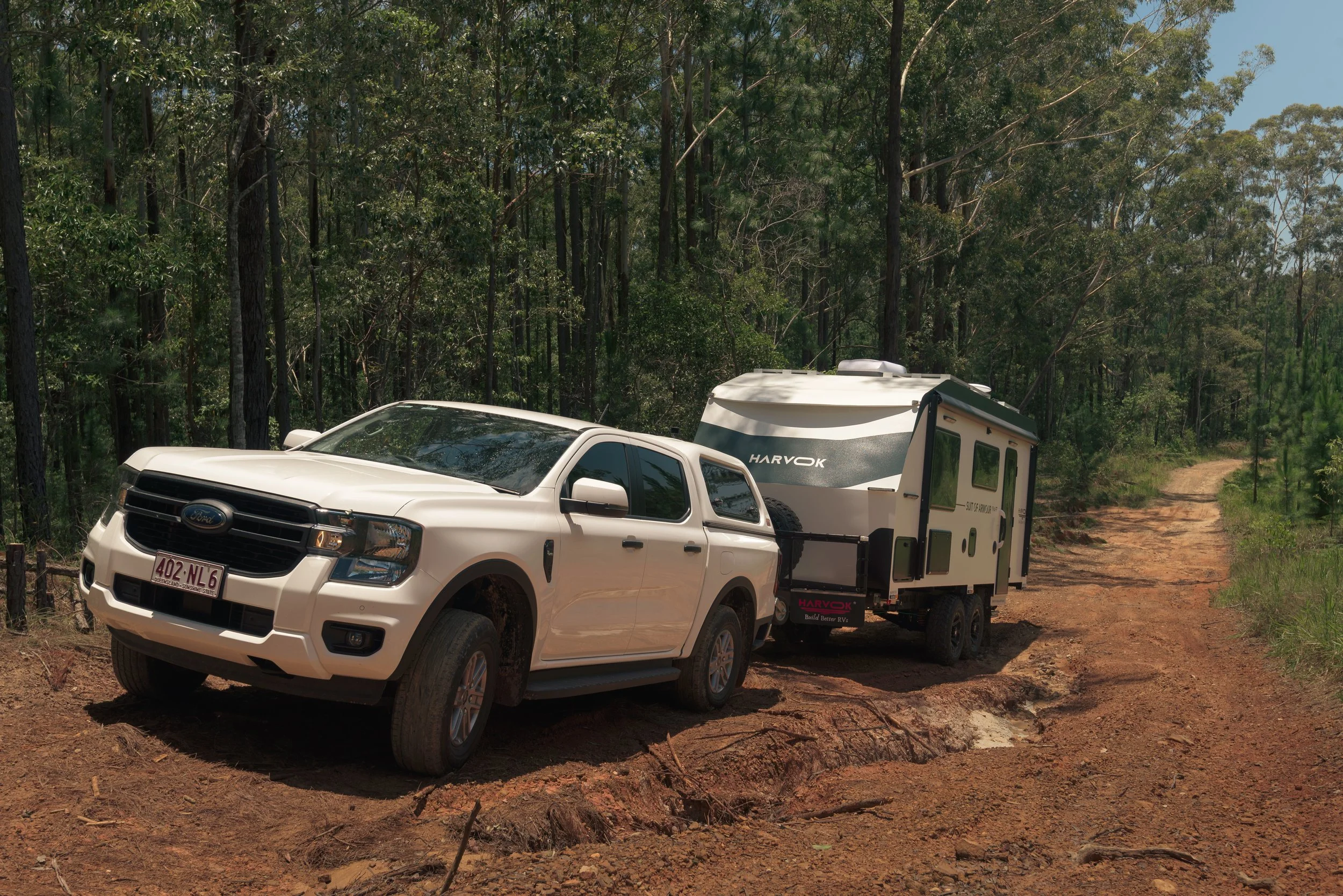 White pickup truck connected to a small travel trailer on a dirt path in a forested area.