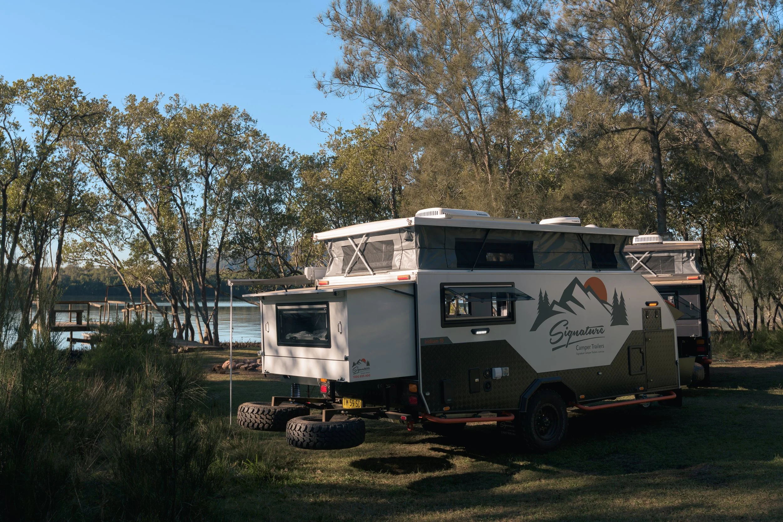 A white camper trailer with mountain and tree graphics parked on grass near a lake, surrounded by trees, under a clear blue sky.
