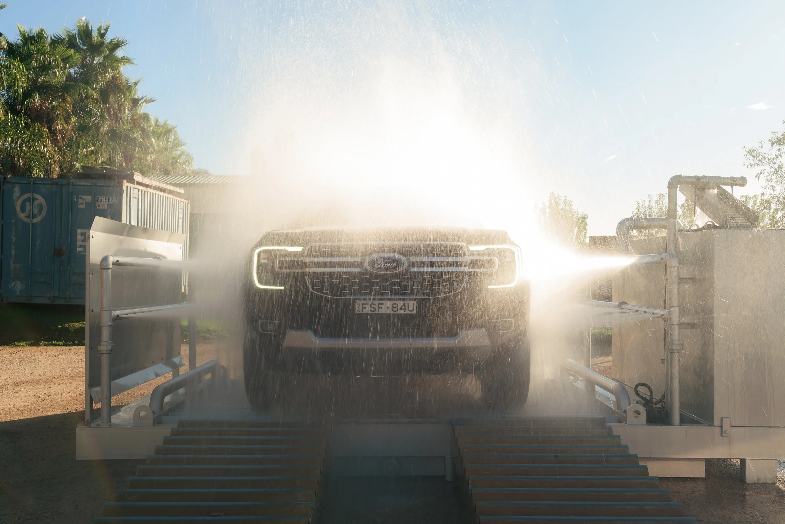 A Ford vehicle is being washed, with water spray and bright sunlight creating a glare. There are trees and a blue sky in the background.