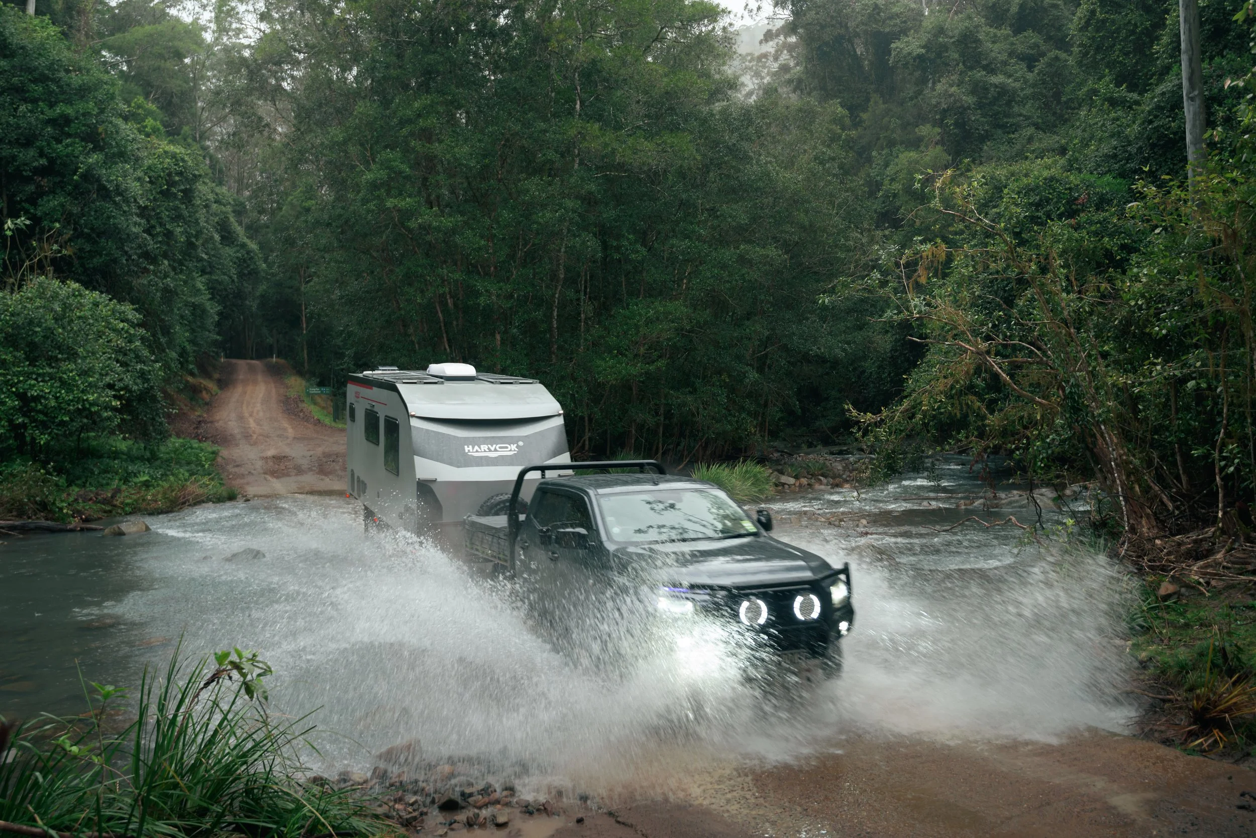 A vehicle crossing a flooded dirt road in a lush green forest during rain, with water splashing around the vehicle.