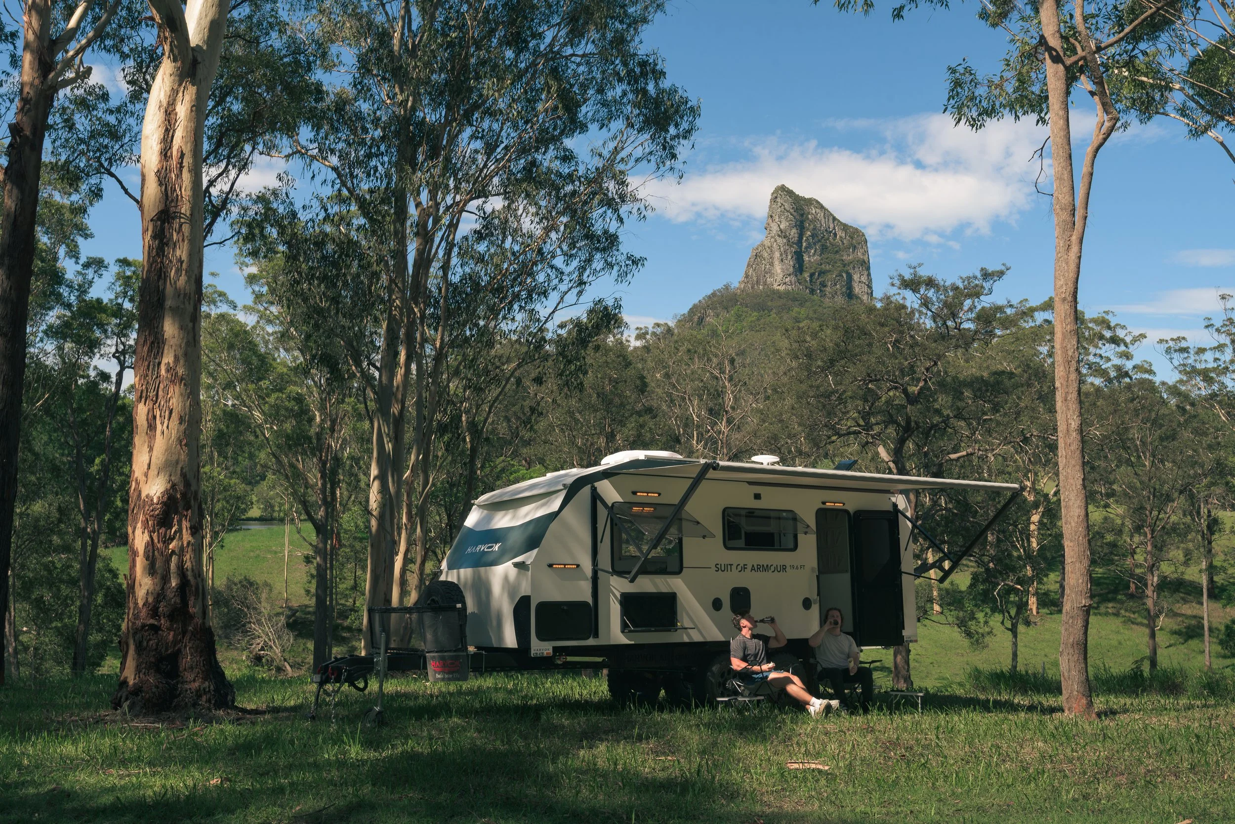 A white camper trailer labeled 'Suit of Armour' parked in a forested area with tall trees and a rock formation in the background. There are two people sitting outside the trailer, one drinking from a cup, and one standing near the trailer. The scene is under a clear blue sky.