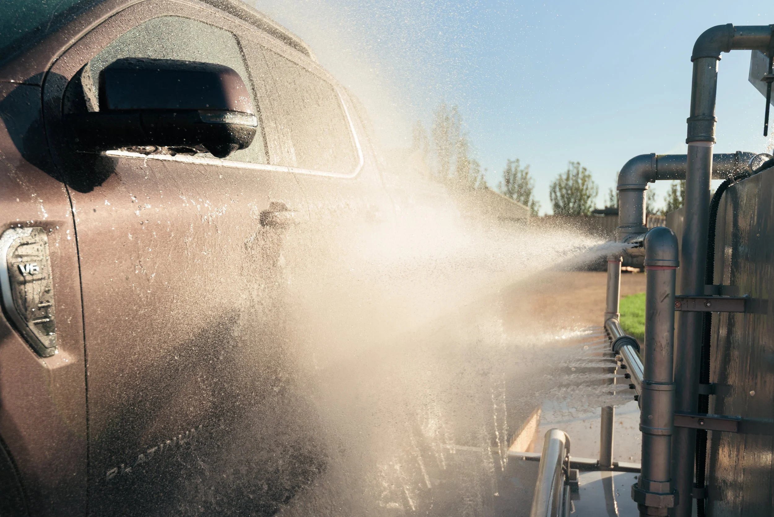 A vehicle being pressure washed at a car wash station under clear blue sky with trees in the background.