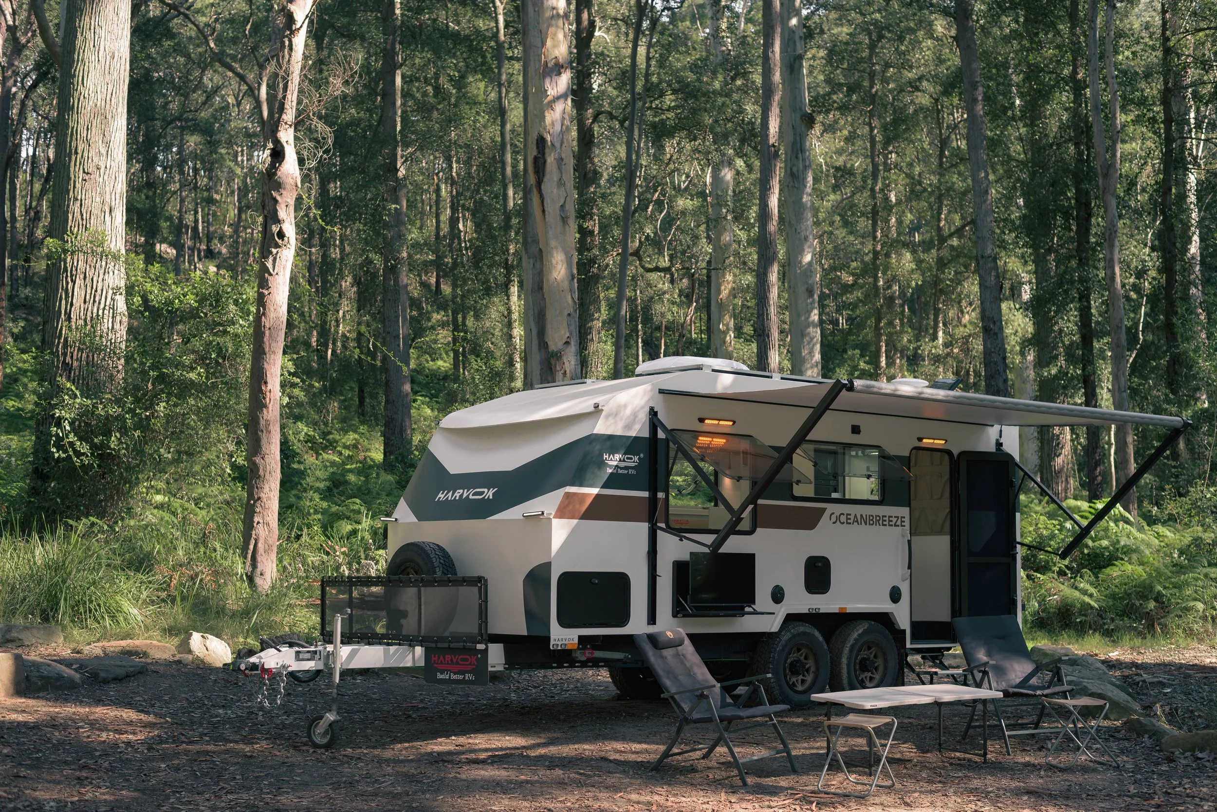 A white travel trailer with black and brown accents parked in a forest clearing surrounded by tall trees. The trailer has an extended awning, outdoor seating, and a small outdoor kitchen area with chairs and a table.