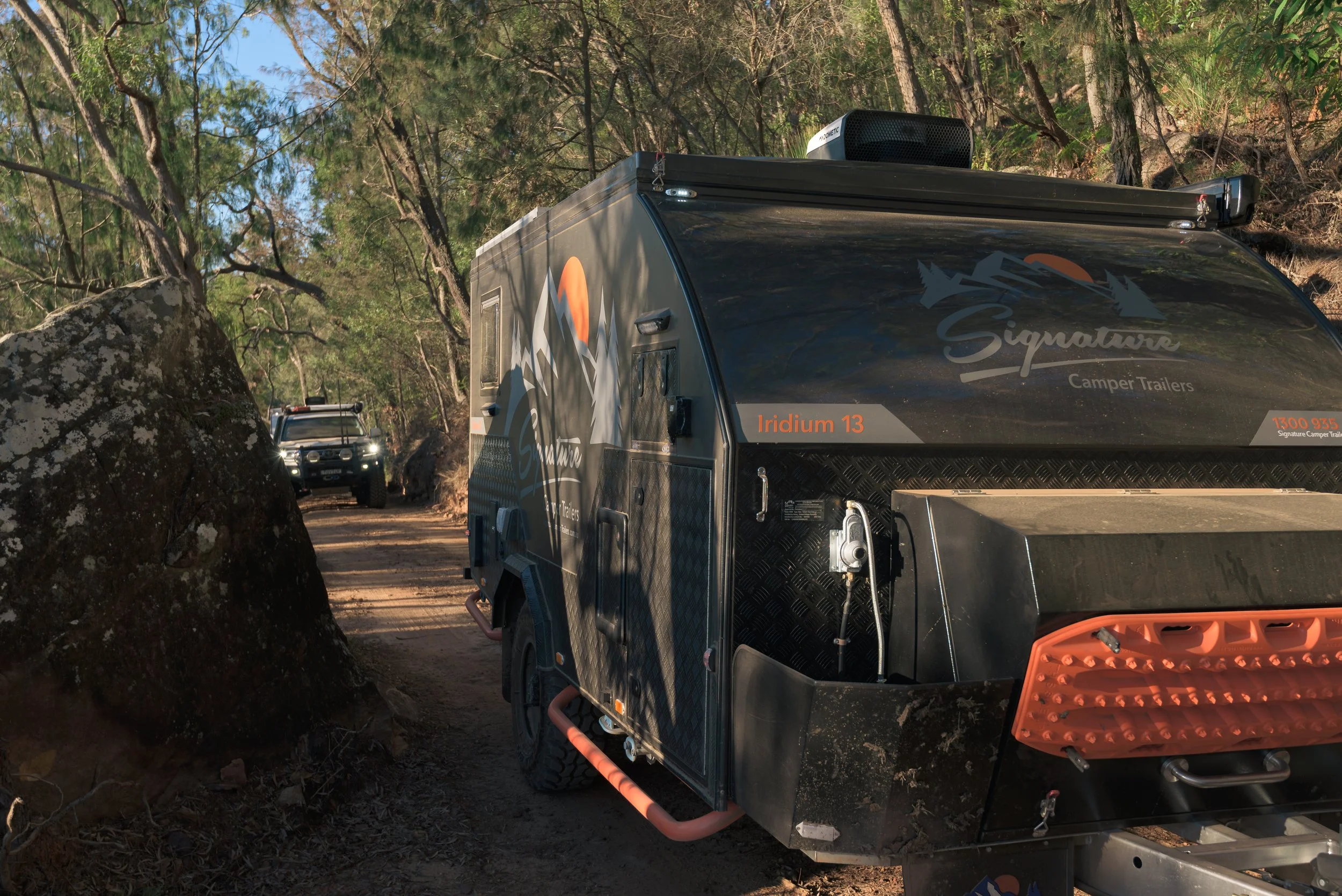 Off-road camper trailer with graphics and a logo on its side, parked along a forest trail with trees and rocks, and a support vehicle further back on the trail.