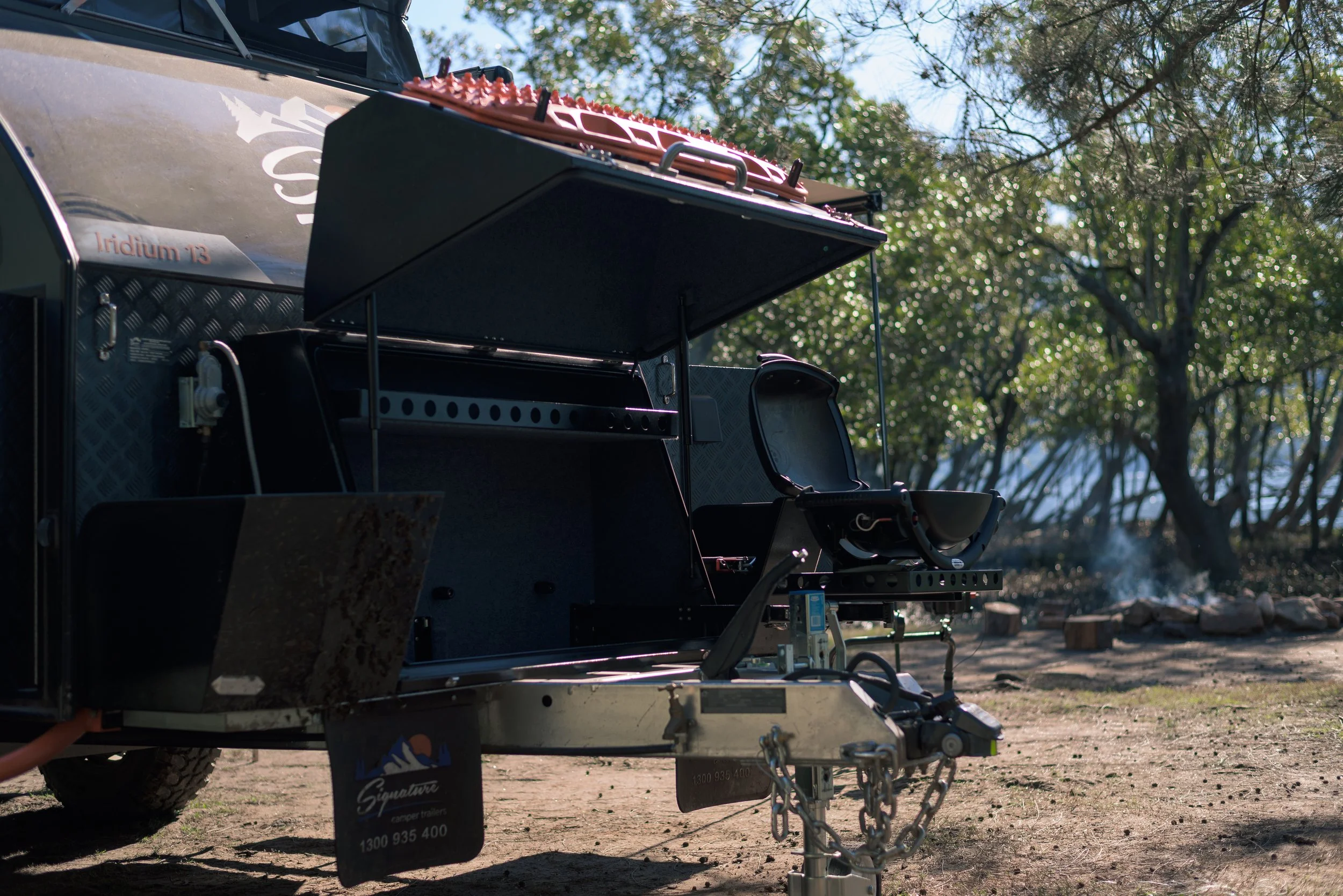 A close-up of a black adventure or camper trailer, focusing on its open side storage compartment and hitch, with a background of trees and a clear sky.