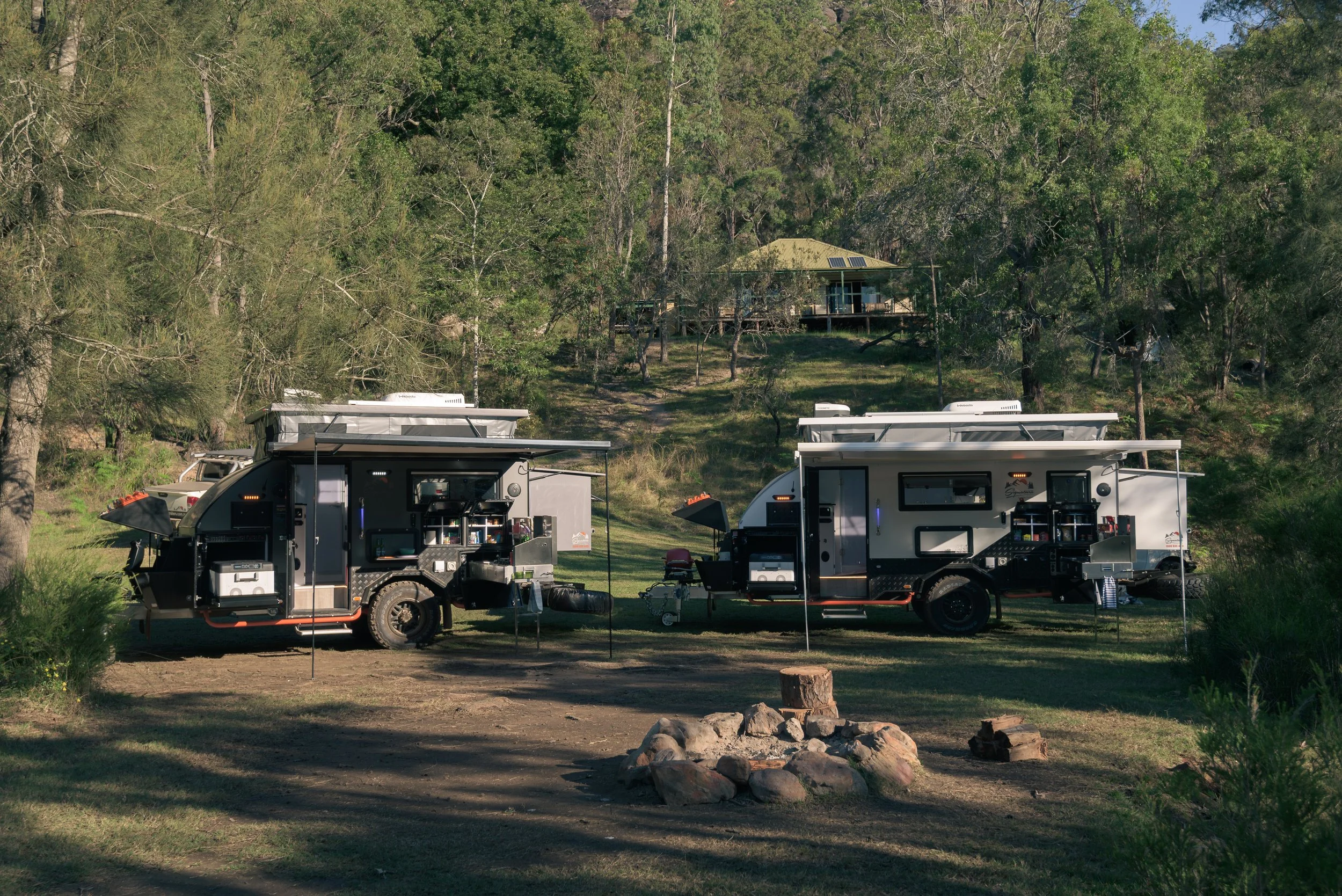 Two camping trailers parked in a wooded area with trees and a house with a green roof in the background. The trailers are equipped with outdoor kitchen setups and camping gear, and are situated on a dirt area with rocks and logs.