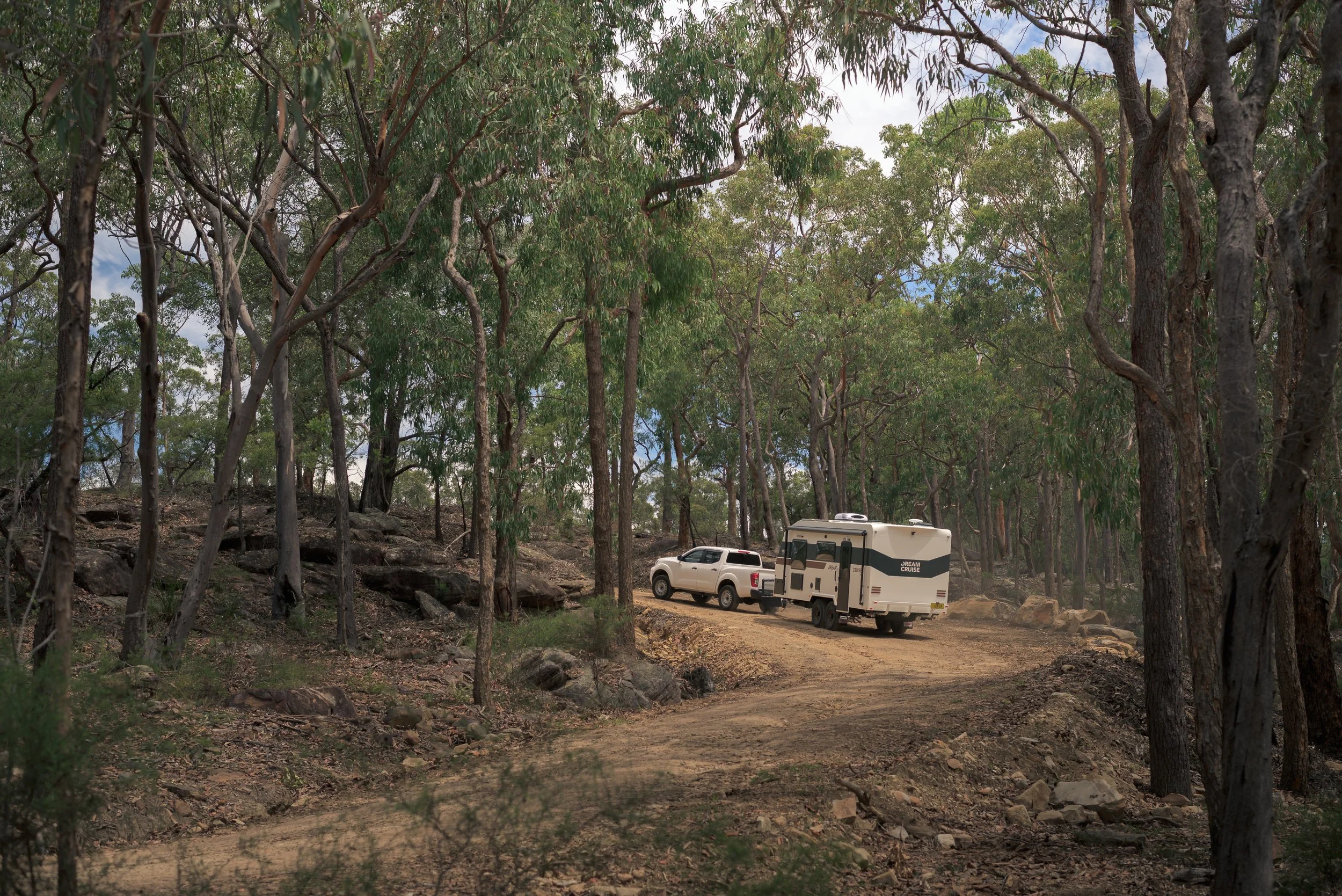 A small white truck towing a white camper trailer on a dirt road in a wooded area with tall trees and rocky terrain.